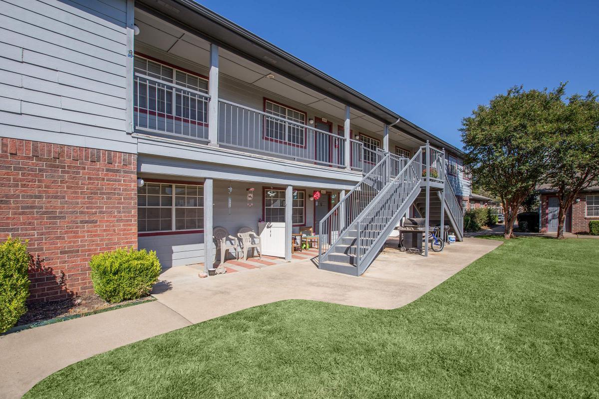 View of a two-story apartment building with a brick and siding exterior. The ground level features a small patio area with chairs, and a staircase leads to the upper level. Neatly trimmed grass and shrubs surround the building, under a clear blue sky.