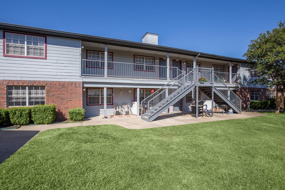 Exterior view of an apartment building featuring two levels, a staircase leading to the upper units, and a neatly maintained lawn. The building has a mix of brick and siding, with several windows and a small seating area visible near the entrance. A bicycle is parked outside.