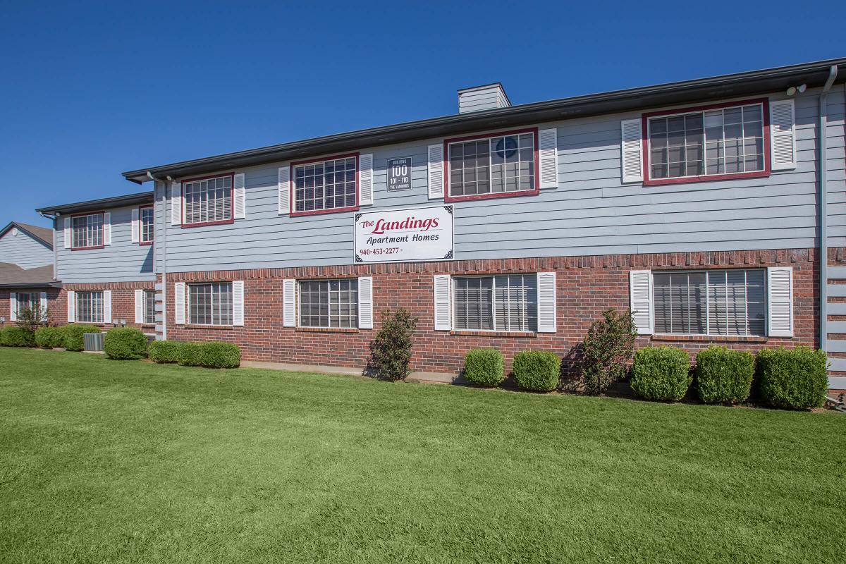 Exterior view of The Landings Apartment Homes, showcasing a two-story building with blue siding and brick accents. A large sign displays the name, and well-maintained green grass and shrubs surround the property under a clear blue sky.