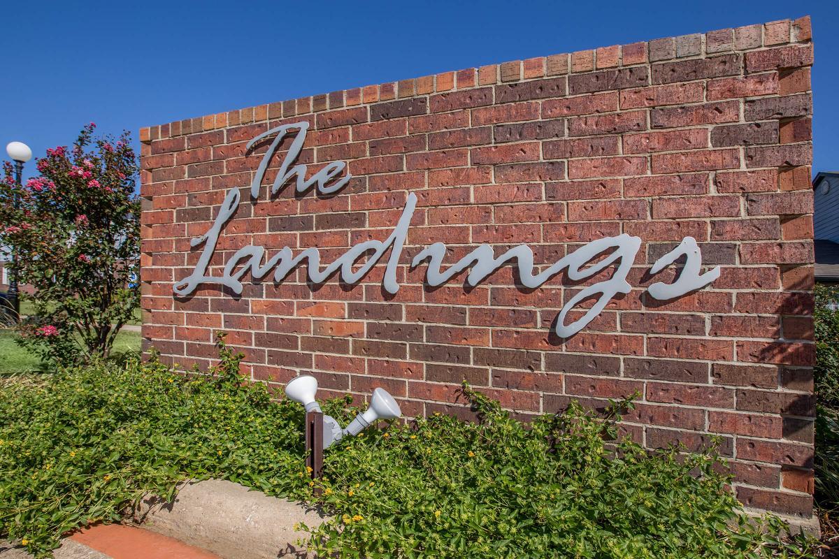 Sign for "The Landings" featuring stylized silver lettering against a textured brick wall, surrounded by green foliage and landscaping. The background is clear blue sky, creating a bright and welcoming atmosphere.