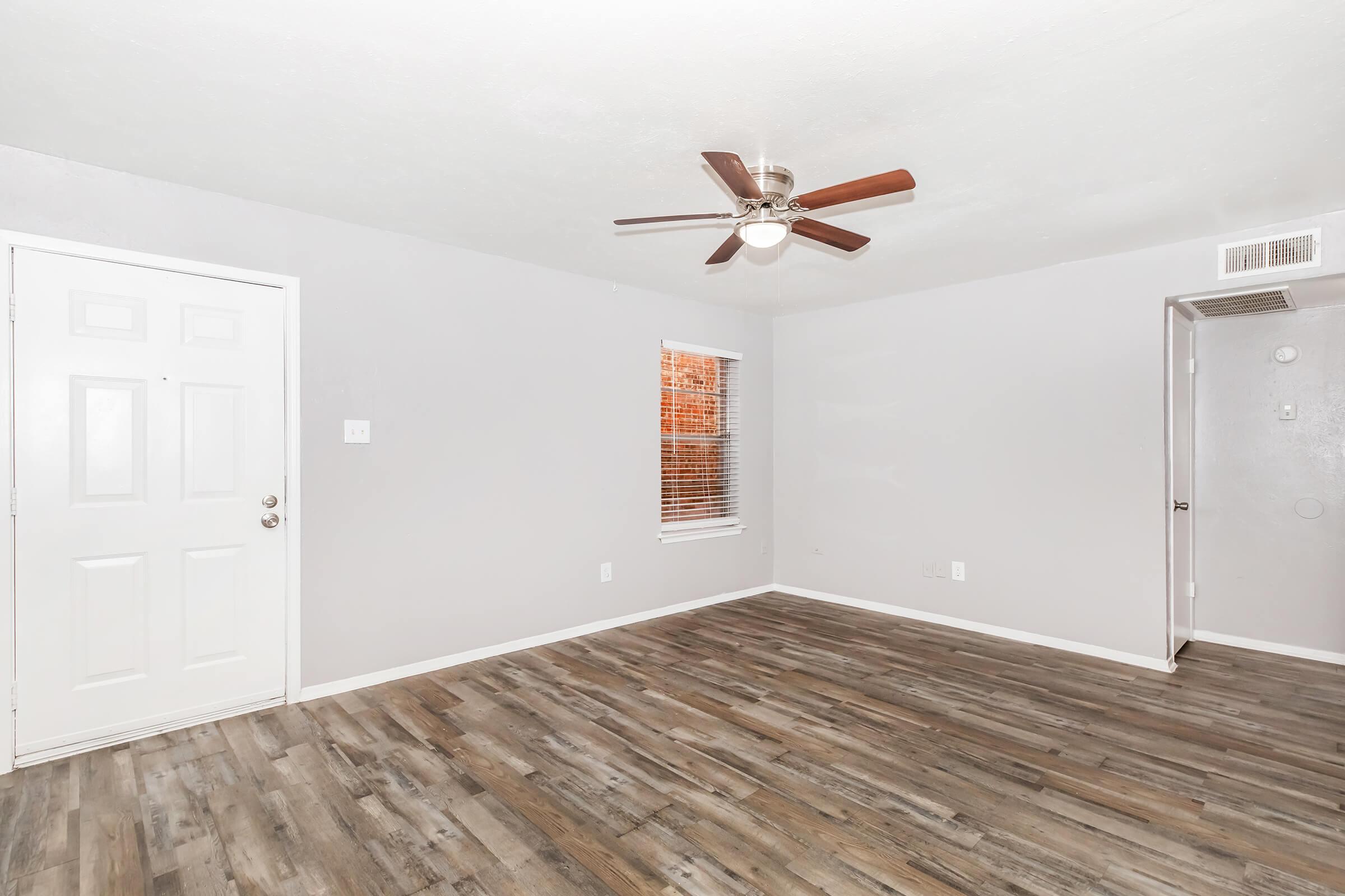 Empty room with gray walls and wooden flooring. It features a ceiling fan with wooden blades, a white door on the left, and a window with blinds allowing natural light. The space is uncluttered and ready for furniture arrangement.