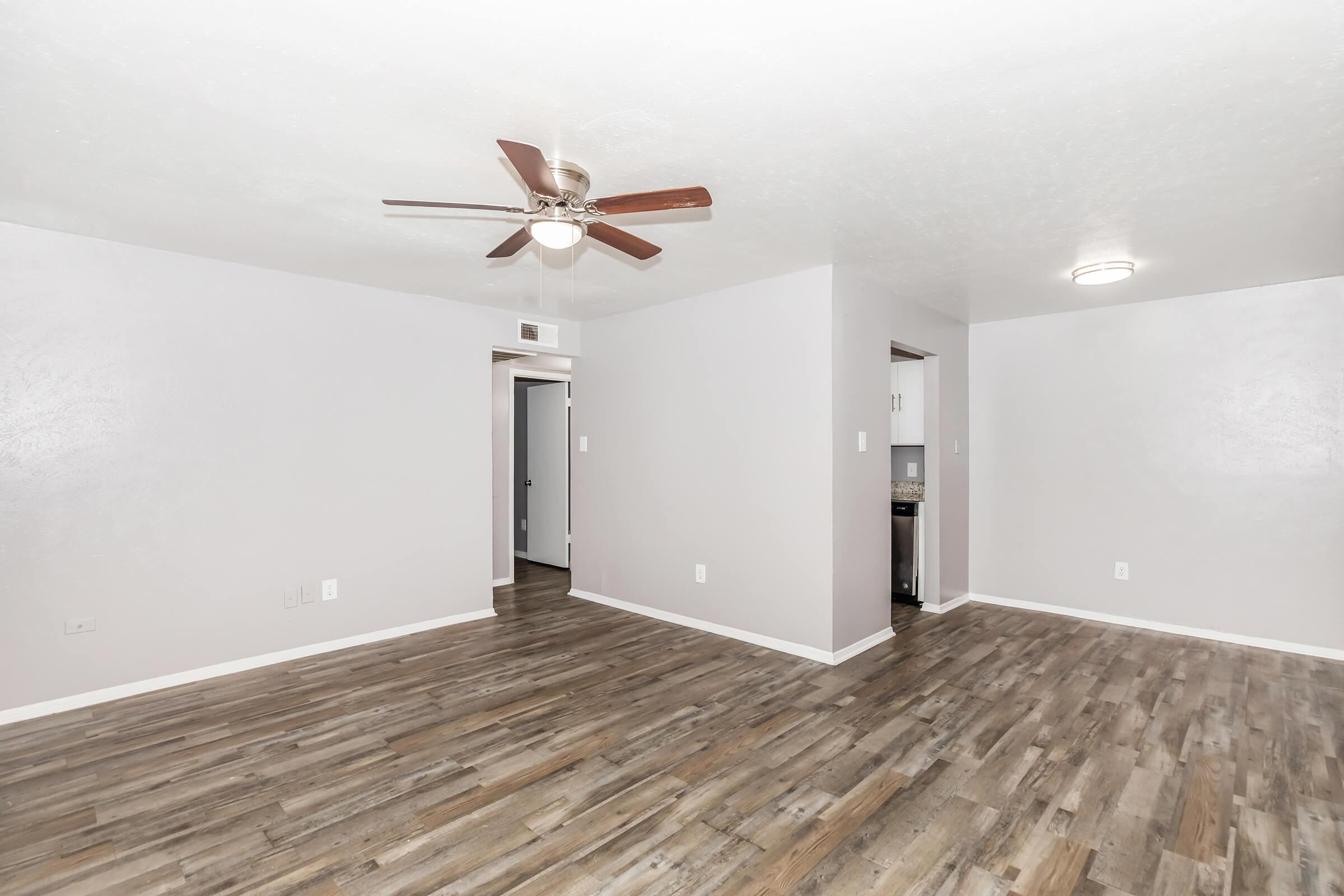 A spacious, lightly colored living room featuring a ceiling fan, new laminate flooring, and white walls. An adjoining doorway leads to another room, while a kitchen area is visible in the background. The overall atmosphere is bright and open, ideal for modern living.