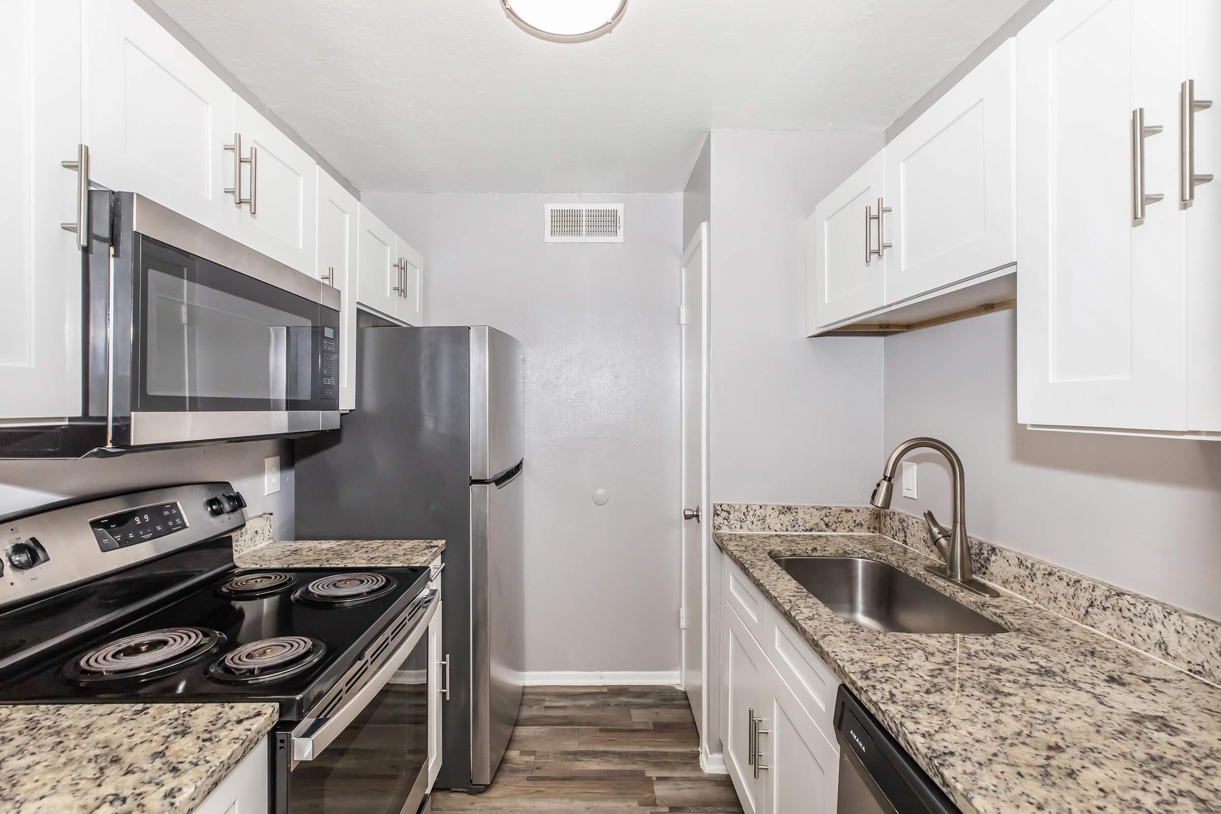 Modern kitchen featuring stainless steel appliances including a microwave, oven, refrigerator, and sleek countertops. White cabinets with contemporary handles provide ample storage. The space is well-lit with neutral walls and a mix of dark and light flooring, creating an inviting atmosphere.