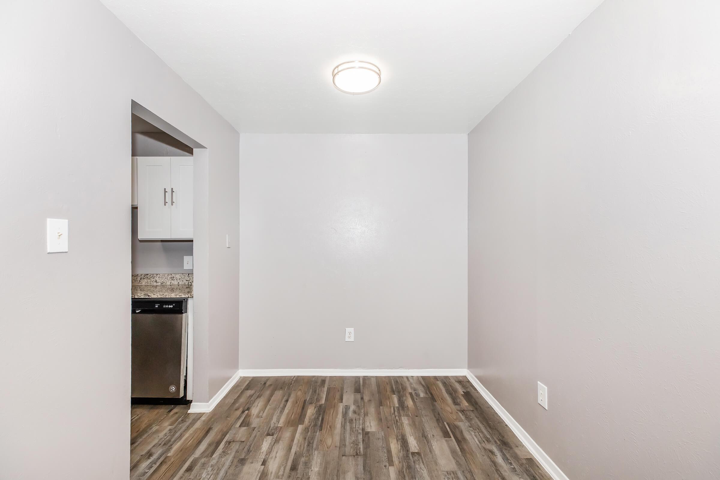 Minimalist interior of a room featuring light gray walls and wood-like flooring. A small opening leads to a kitchen, visible appliances partially seen. The ceiling has a central light fixture, adding illumination to the space. The overall ambiance is modern and uncluttered.
