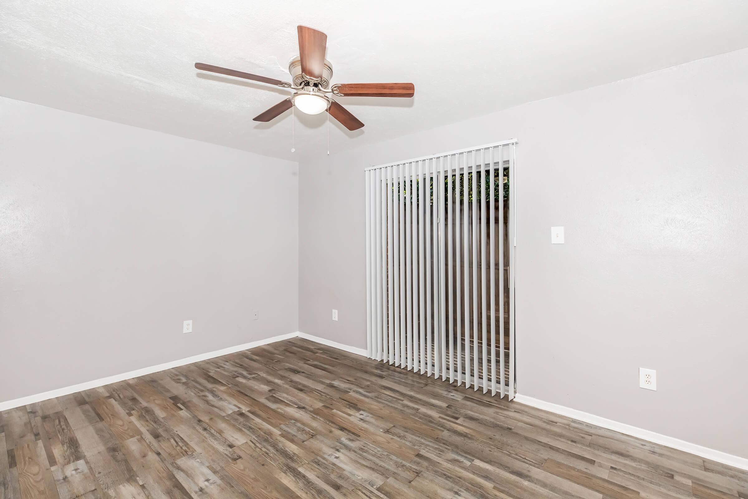 A light gray room with wooden flooring and a ceiling fan featuring wooden blades. The room has vertical blinds covering a window, allowing some natural light. The walls are bare, creating a clean and minimalist look. There are no furniture or decor items visible in the space.