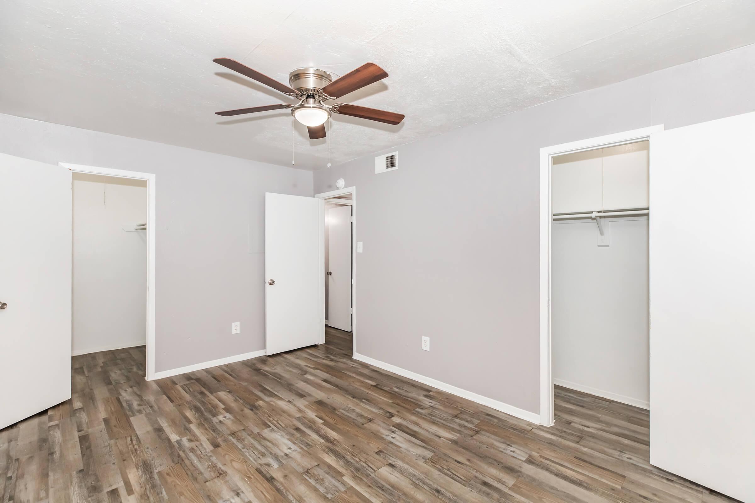 A small, empty room with gray walls and a wooden floor. It features a ceiling fan and three doors leading to closets or other areas, creating an open, airy feel. The space is well-lit and has a clean, modern appearance, suitable for various purposes such as a bedroom or office.