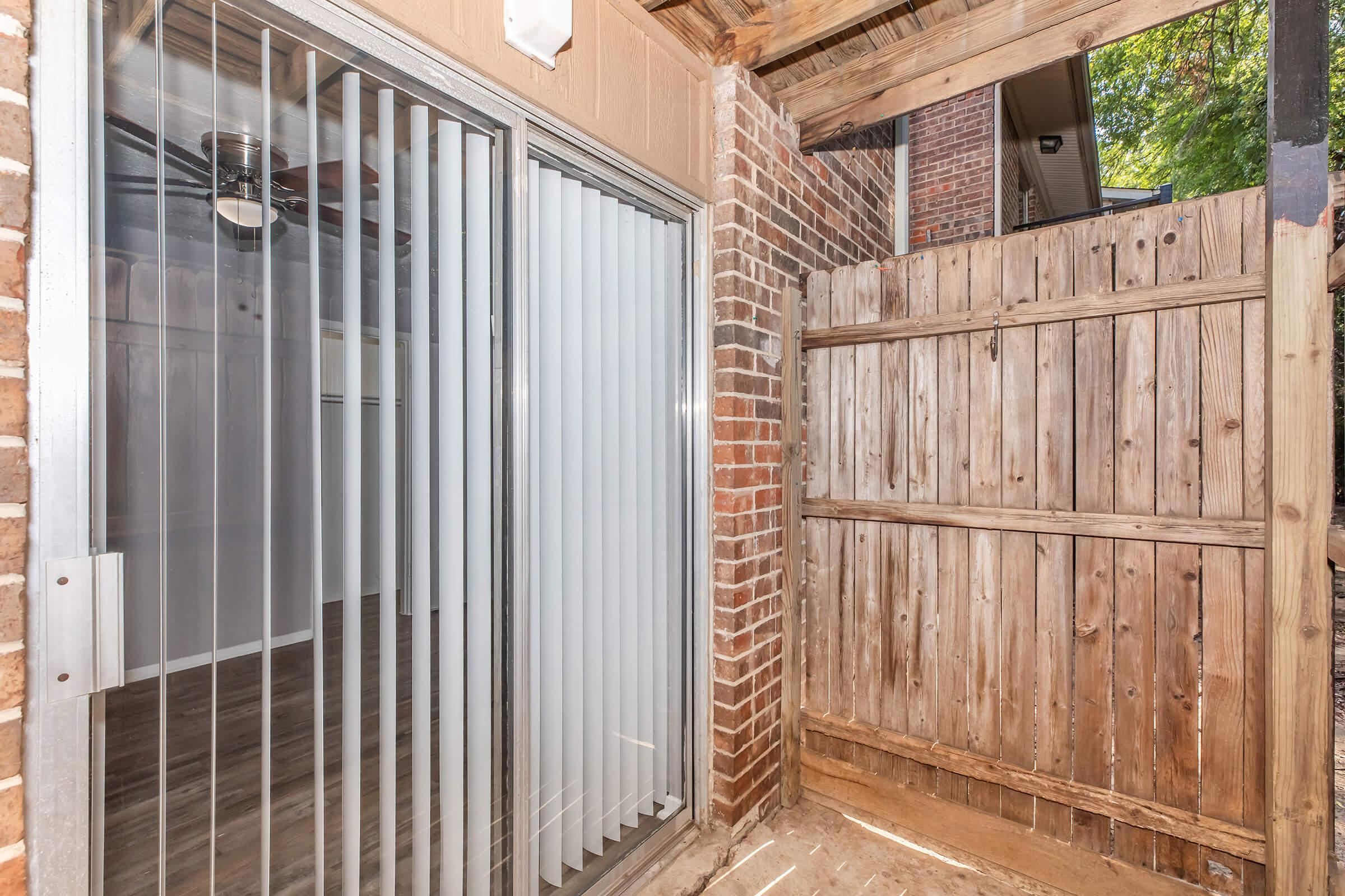 A view of a small outdoor area featuring a sliding glass door with vertical blinds, brick walls, and a wooden fence. The space is well-lit and includes a roof overhang. The flooring appears to be concrete, and there are wooden beams supporting the structure above.