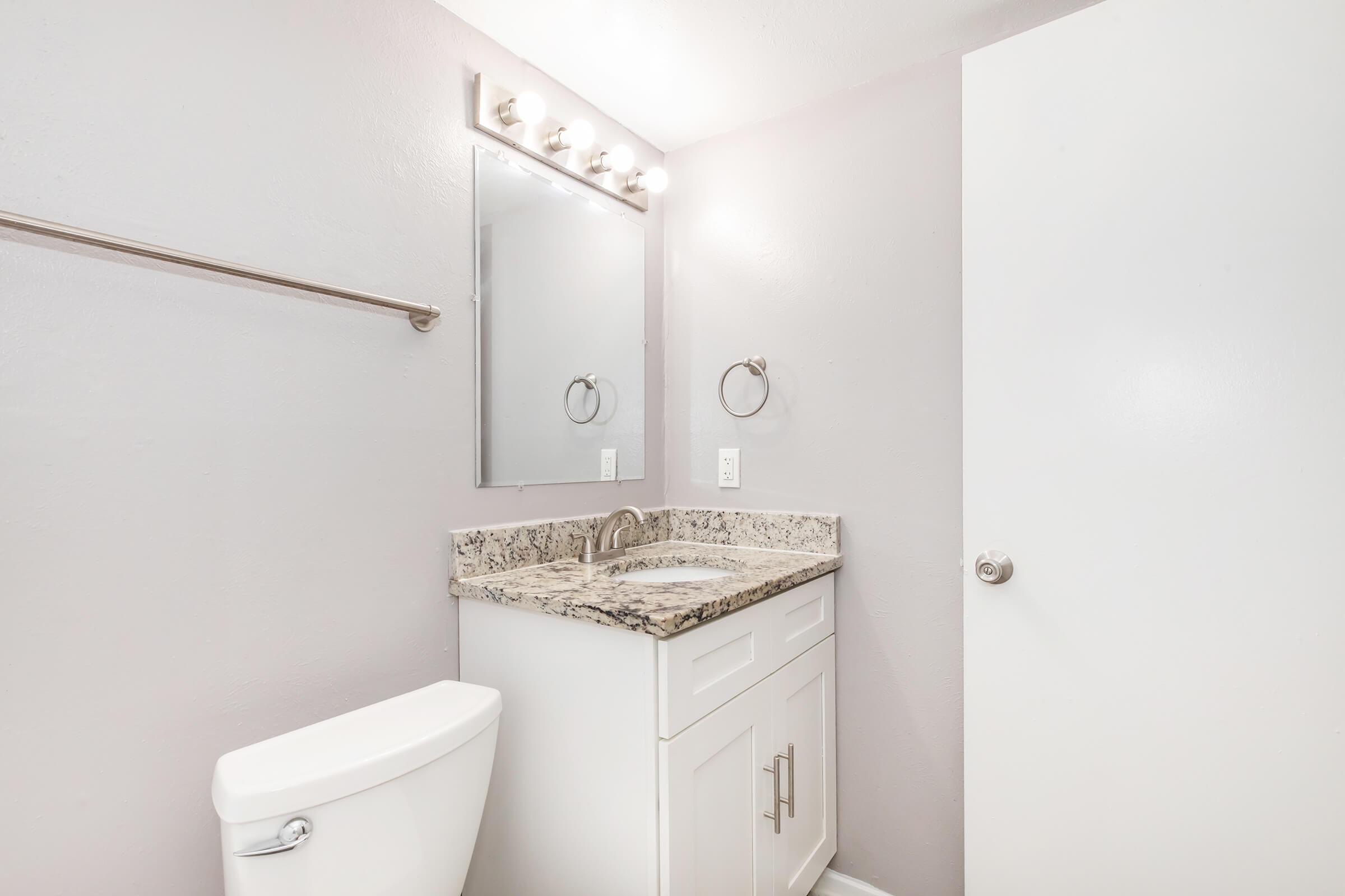 A modern bathroom featuring a light gray wall, a granite countertop with a sink, a mirror with overhead lights, a towel ring, and a white toilet. The bathroom door is partially open, revealing a clean and organized space.