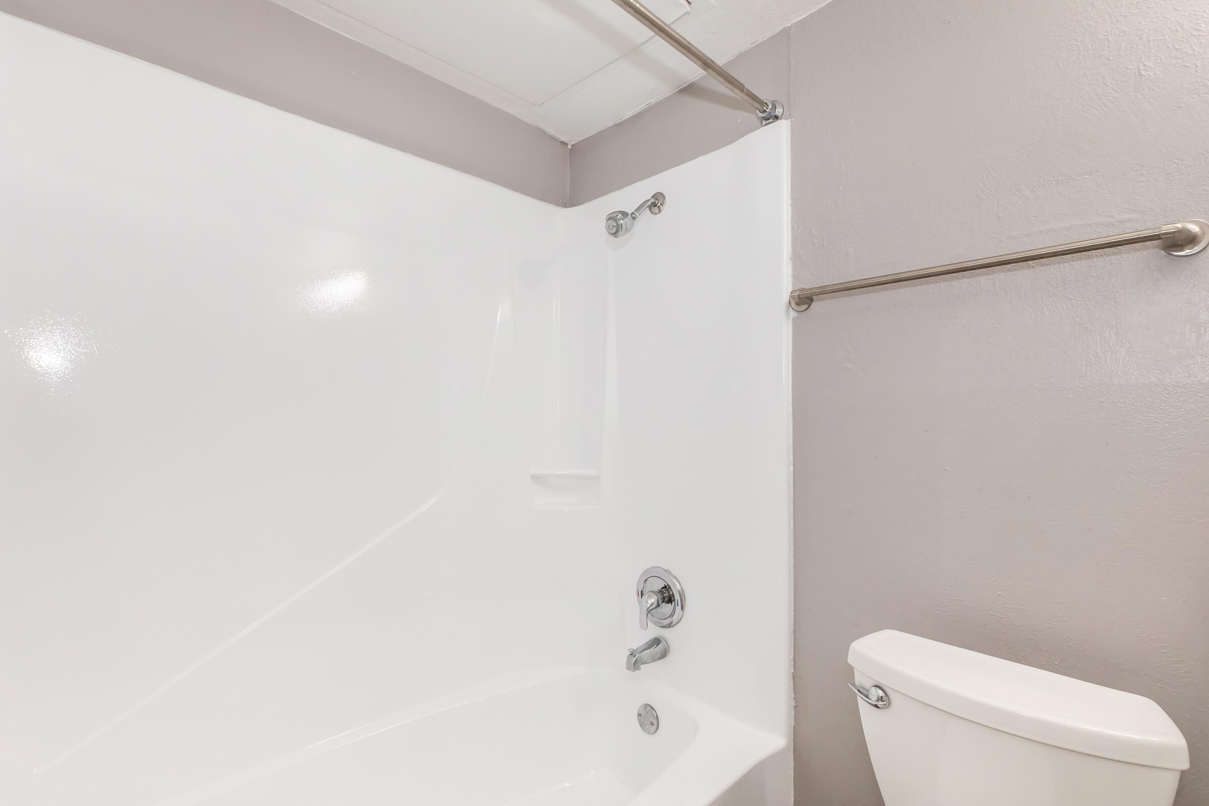 A clean, modern bathroom featuring a white bathtub with a showerhead, a wall-mounted towel bar, and a toilet. The walls are painted in a light purple color, creating a fresh and inviting atmosphere. The overall design is simple and functional, ideal for a contemporary space.