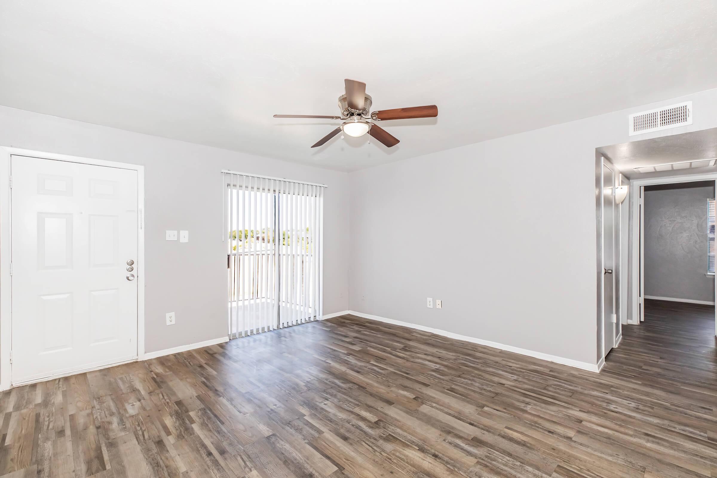 Interior view of a bright, empty living room featuring a ceiling fan, laminate flooring, and a large window with vertical blinds. A door leads outside, and there are doorways to other rooms in the background. The walls are painted in a light gray color, creating an airy atmosphere.