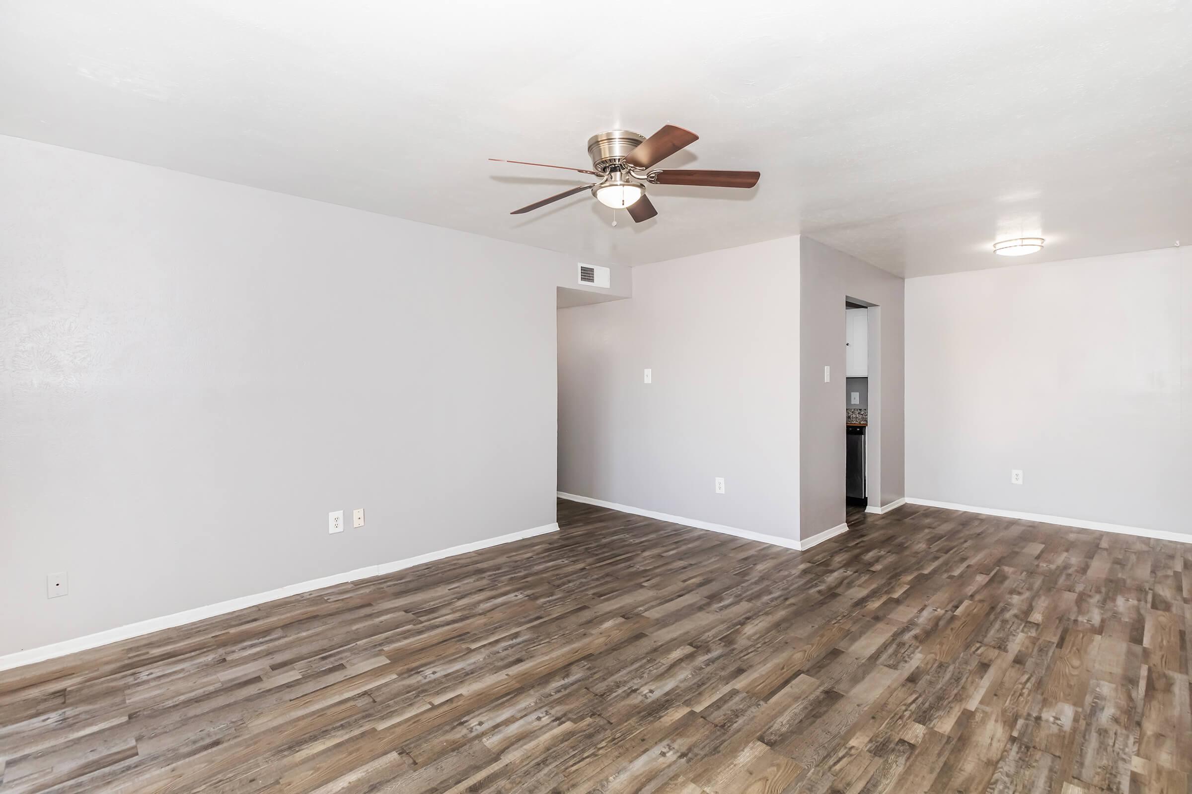A spacious interior living area featuring light gray walls, a ceiling fan, and wooden flooring. The room is empty with a few visible light fixtures and an open entryway into another room, creating a bright and airy atmosphere.