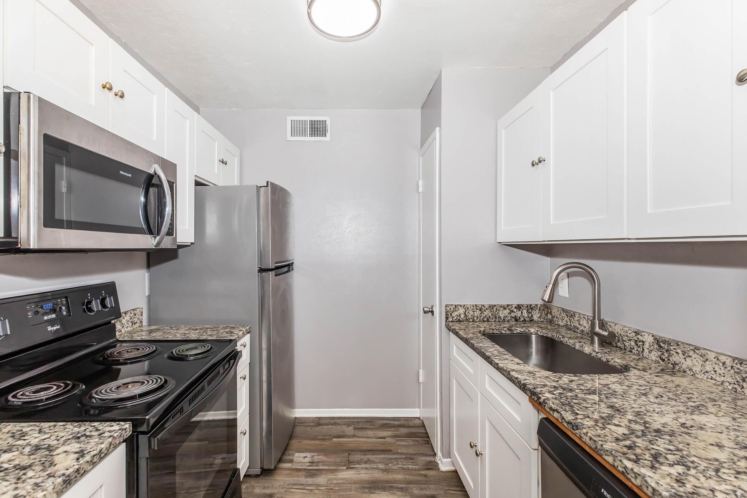 A modern kitchen featuring white cabinets, a stainless steel refrigerator, a microwave, and an oven. The countertops are made of speckled granite, and there is a single basin sink. The walls are painted light gray, and the flooring appears to be laminate. A ceiling light fixture illuminates the space.