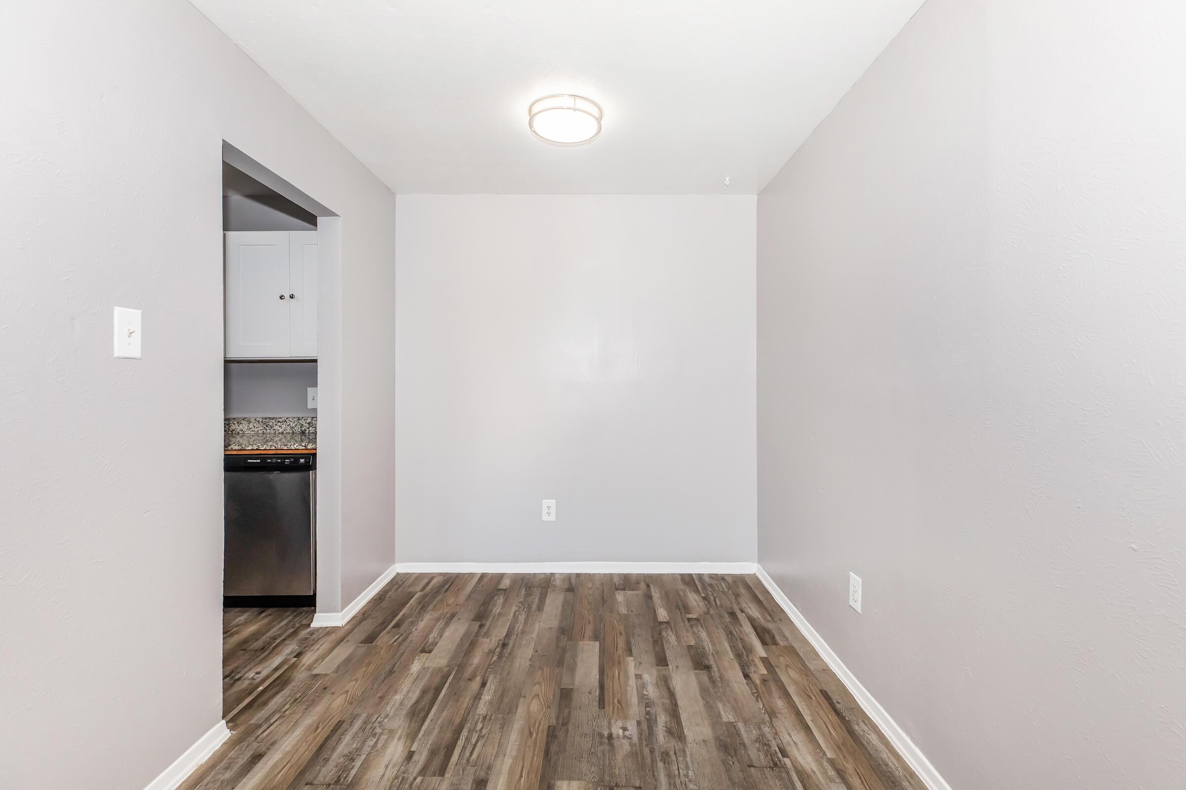 A simple, empty room with light gray walls and a modern ceiling light. The floor is covered in wood-like laminate. On one side, there's an open doorway leading to a kitchen area with white cabinets and a stainless steel appliance visible. The space feels clean and ready for furnishing.