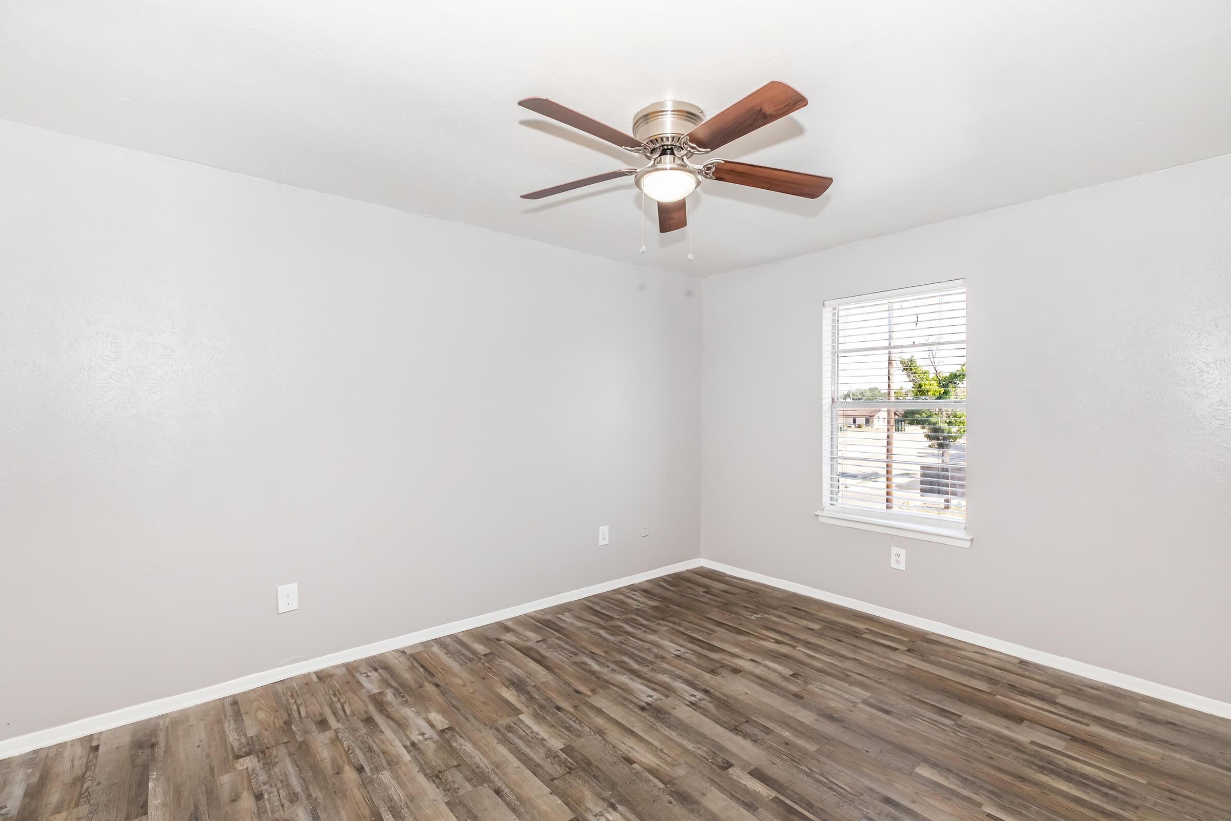 Empty room with light gray walls and wooden laminate flooring. A ceiling fan with wooden blades is mounted in the center. A window with horizontal blinds allows natural light to enter, and the view outside shows greenery. The space is clean and minimalistic, suitable for various furnishing options.