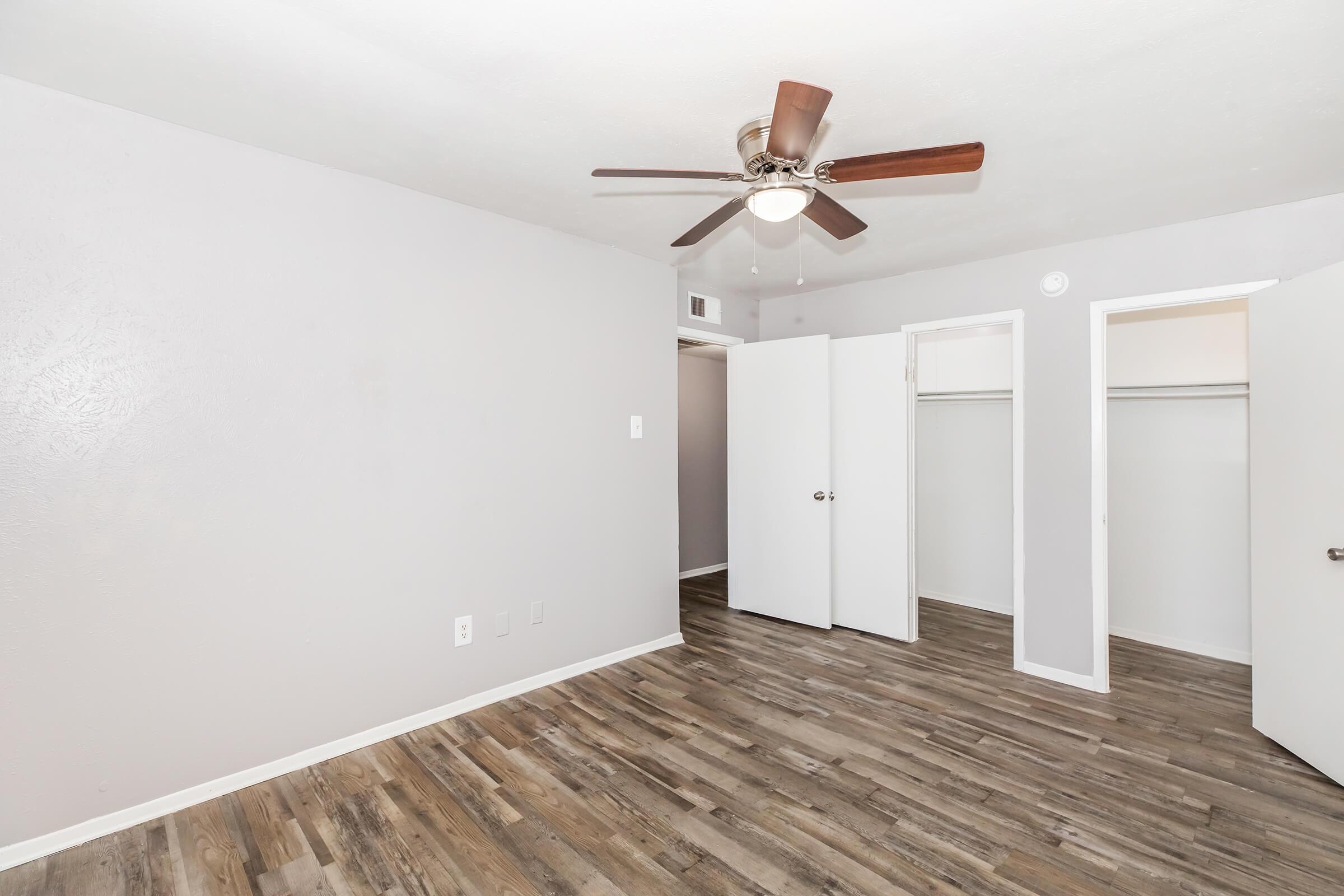 A spacious room featuring light gray walls and wood-like flooring. There is a ceiling fan with wooden blades, and two white closet doors on the right. The room has a clean and minimalist design, with ample natural light and a neutral color palette, creating a bright and inviting atmosphere.