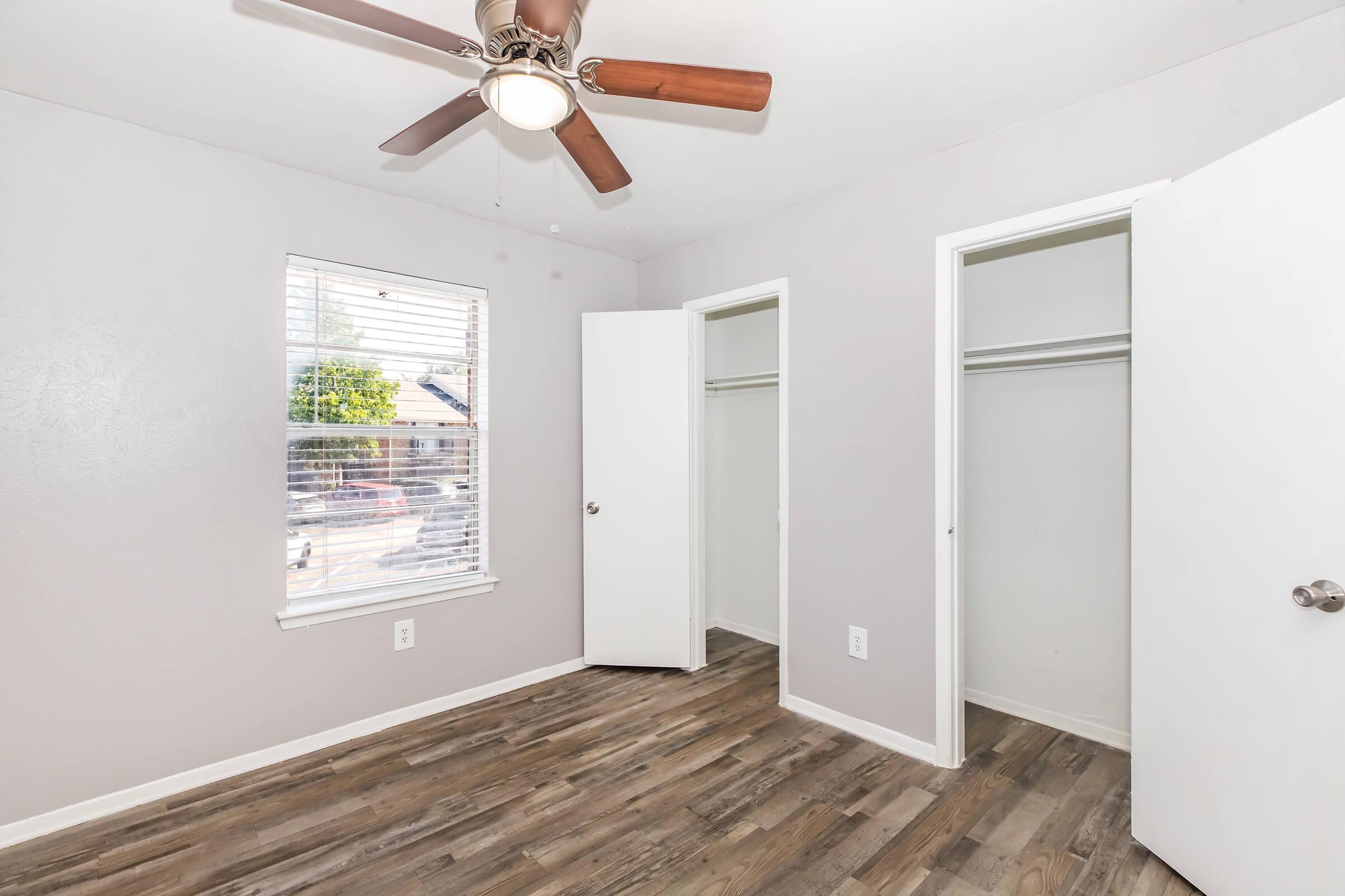 A well-lit bedroom featuring a ceiling fan, light gray walls, and wooden flooring. There is a window with blinds allowing natural light, along with two white closet doors on opposite walls, providing storage space. The room is empty and appears freshly painted and modern.
