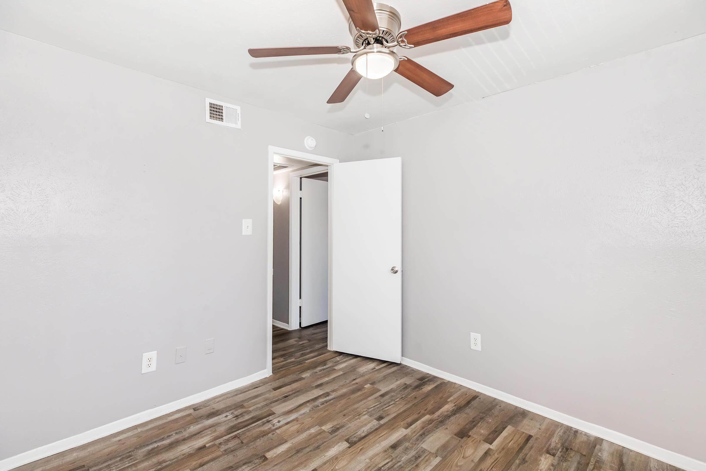 A well-lit, neutral-colored room featuring a ceiling fan and a door partially open to another space. The floor is covered with wood-like laminate, and the walls are painted light gray. Ideal for a bedroom or office, the decor is simple and modern.