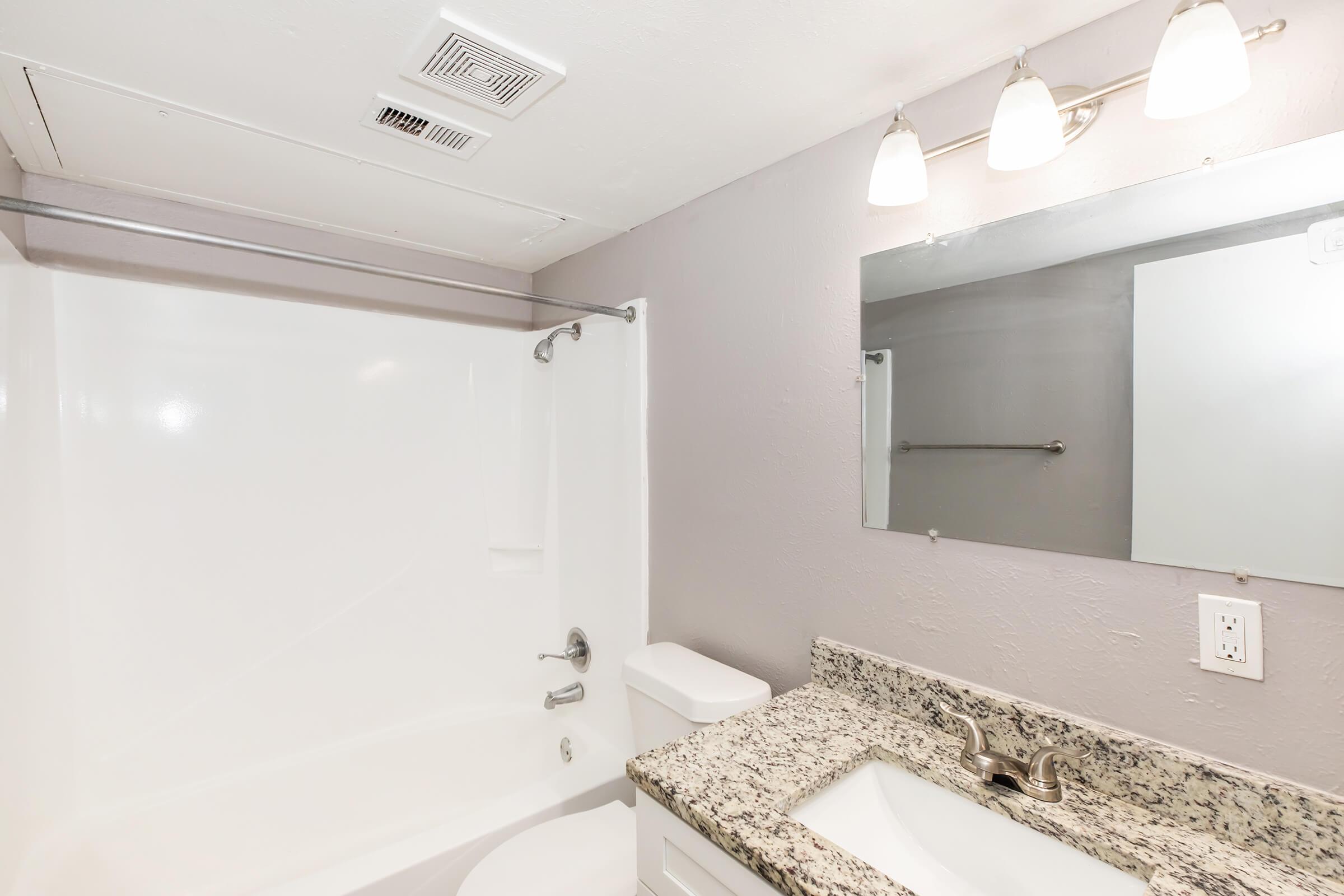 A clean and modern bathroom featuring a white bathtub with a shower, a granite countertop with a sink, and a large mirror above the sink. The walls are painted a light lavender color, and there are three light fixtures above the mirror, providing ample lighting.