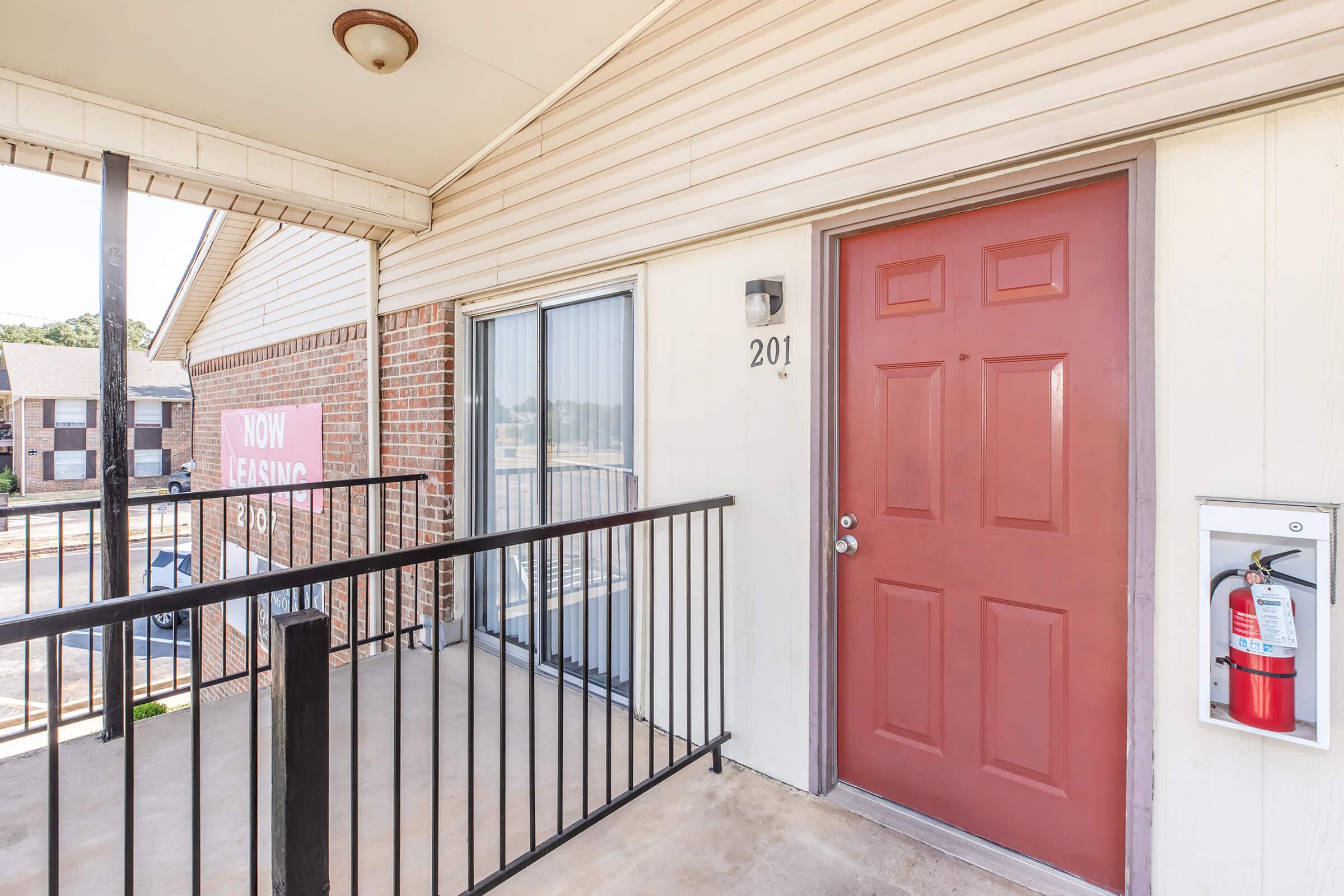 A view of an apartment door labeled "201" on a balcony. The door is red, flanked by a window on the left, and there is a fire extinguisher mounted on the right side of the entrance. A sign in the background reads "NOW LEASING," with a residential building visible beyond the balcony.