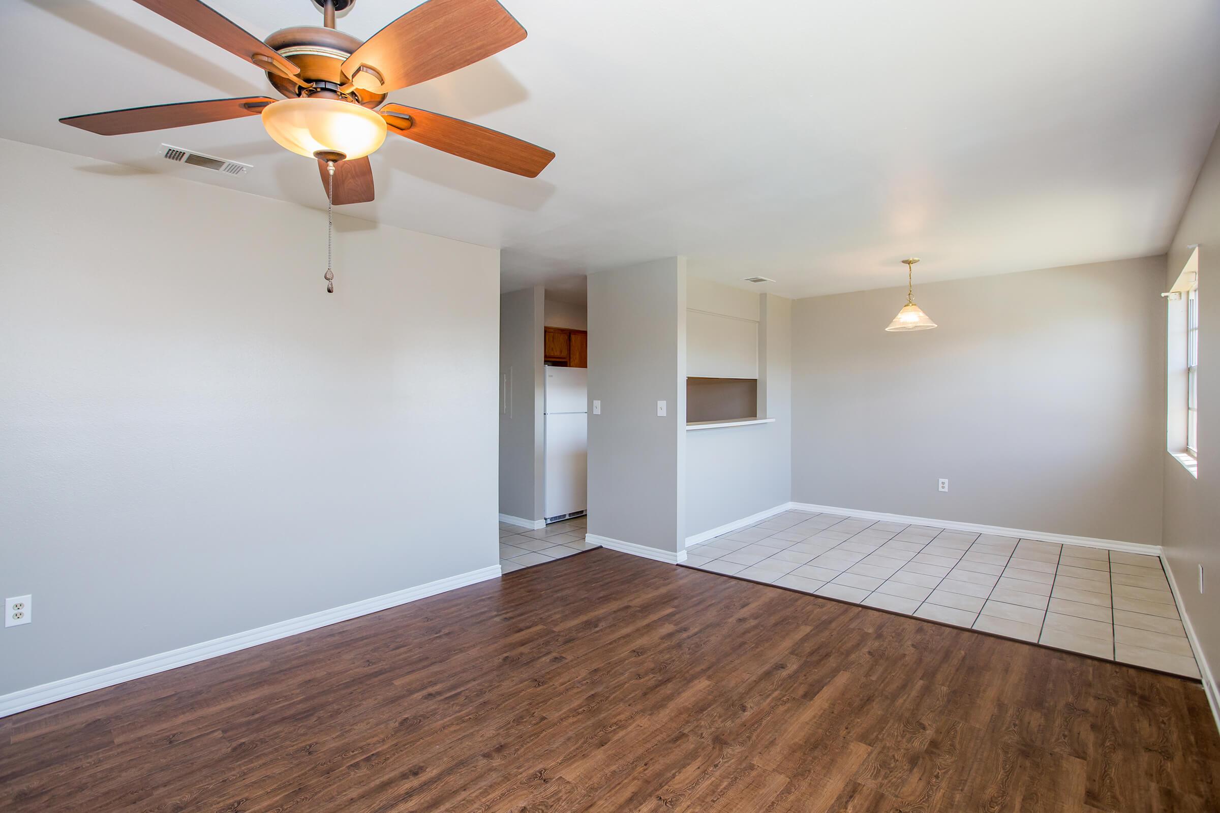 Spacious living area with hardwood flooring, a ceiling fan, and a neutral color palette. Adjacent to a kitchen visible through a pass-through opening. Natural light coming through a window on the right, and a tile-floored dining area in the background, enhancing the open layout feel.