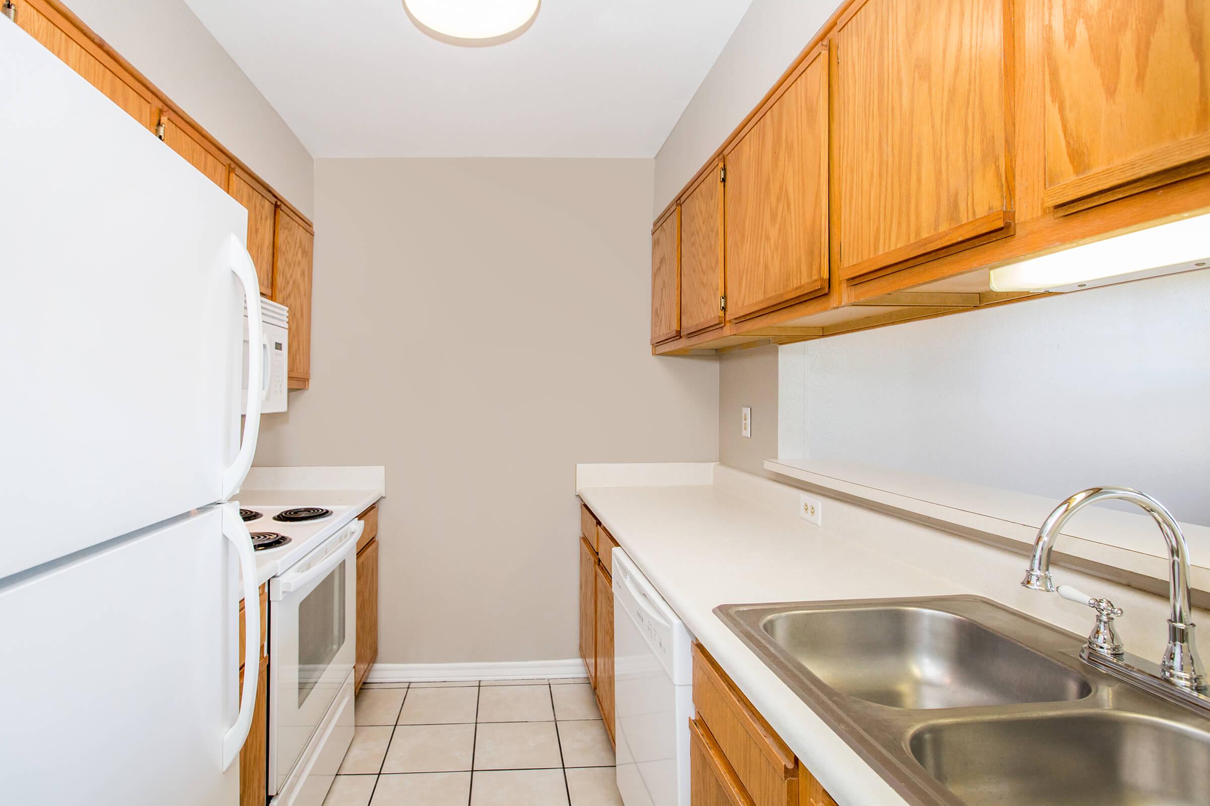 A compact kitchen featuring light wood cabinetry, white appliances including a refrigerator and oven, a double sink, and beige walls. The countertops are beige, and the floor is tiled. Good lighting from a ceiling fixture.
