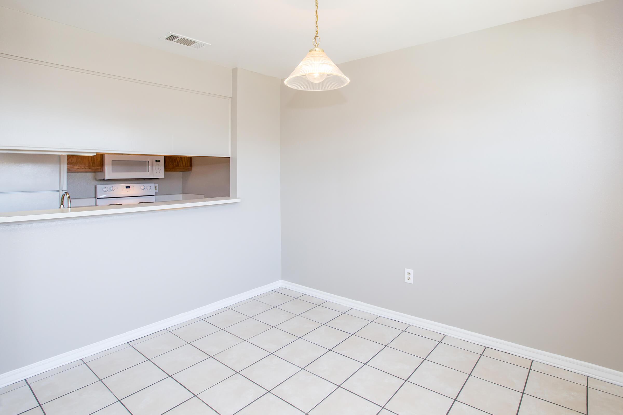 A spacious, empty room with beige walls and tiled flooring. A small kitchen space is visible with a microwave in the background and a pendant light hanging from the ceiling. The area is well-lit, creating a bright and open atmosphere, suitable for a dining or living space.