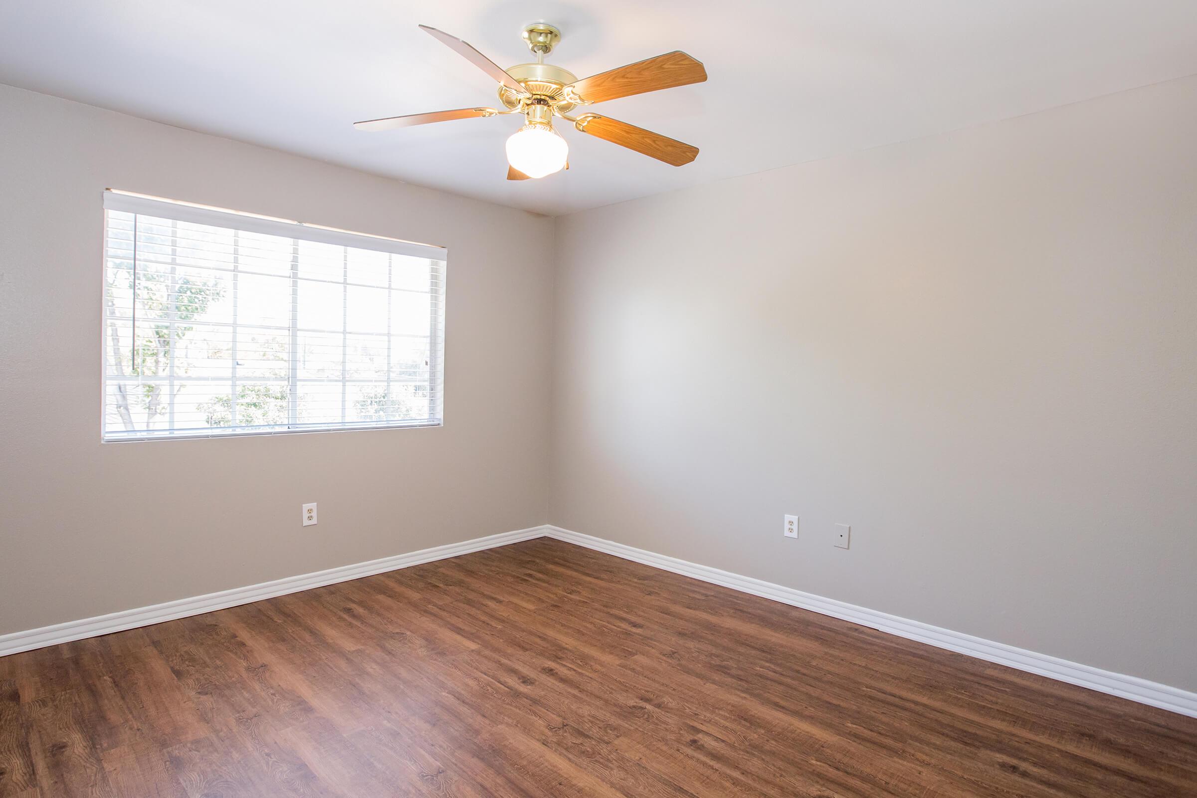 A well-lit empty room featuring light-colored walls and a wooden floor. A ceiling fan with wooden blades hangs from the ceiling, and a window with blinds allows natural light to enter the space. The room appears spacious and ready for furnishing.