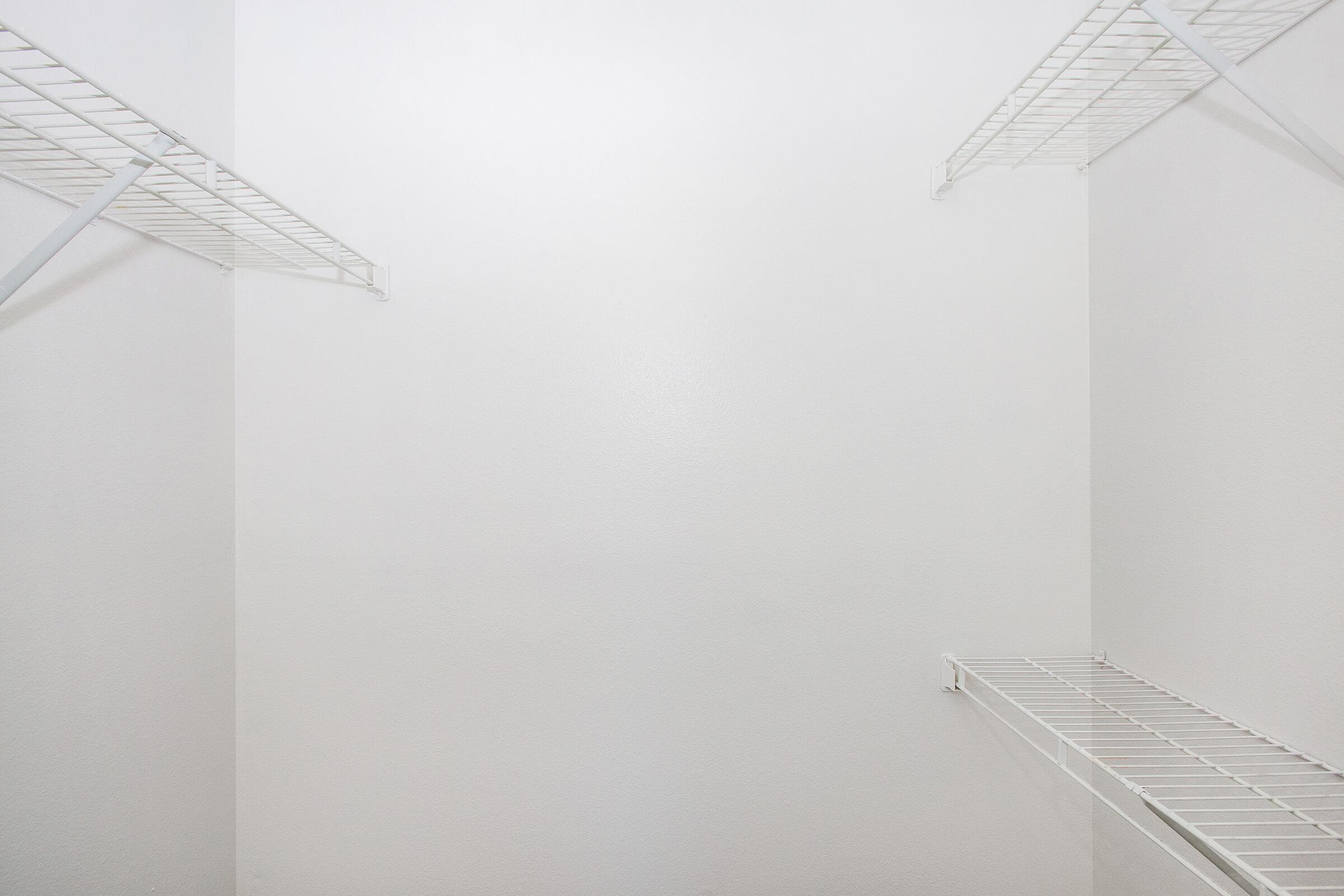 Empty interior of a closet featuring two white wire shelves on opposite walls, with a plain white background. The space appears well-lit and minimalist, emphasizing the lack of contents.