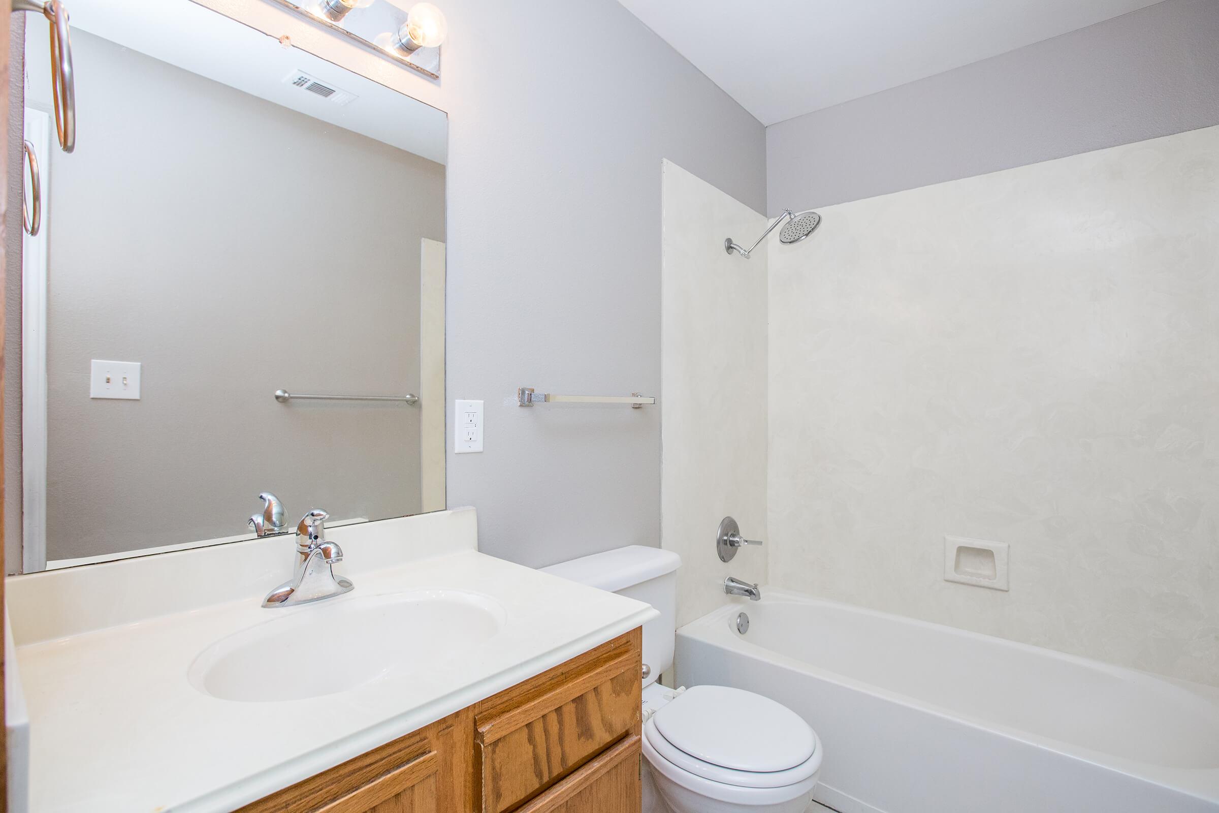 A clean and modern bathroom featuring a white bathtub, a simple sink with wooden cabinetry, and a mirror above the sink. The walls are painted light gray, and there is a shower head above the bathtub. A towel rack is mounted to the left, and the overall space appears well-maintained and inviting.