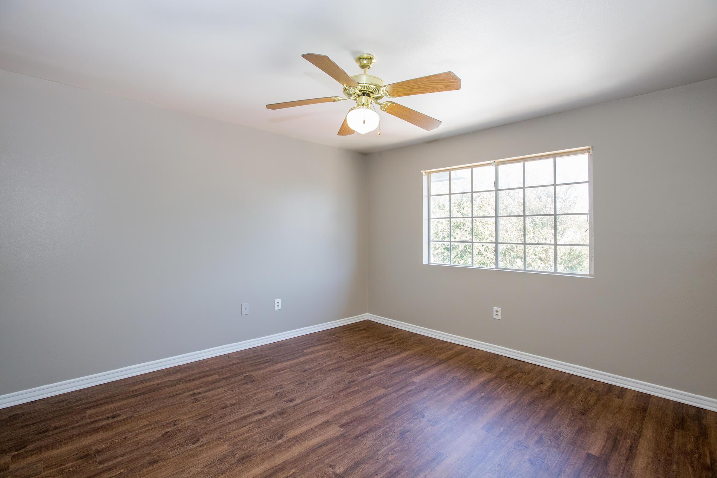 A bright, empty room with light gray walls and a wooden floor. A ceiling fan with wooden blades hangs from the ceiling. A large window with multiple panes allows natural light to enter, illuminating the space. The room is unfurnished, giving it a spacious and open feel.