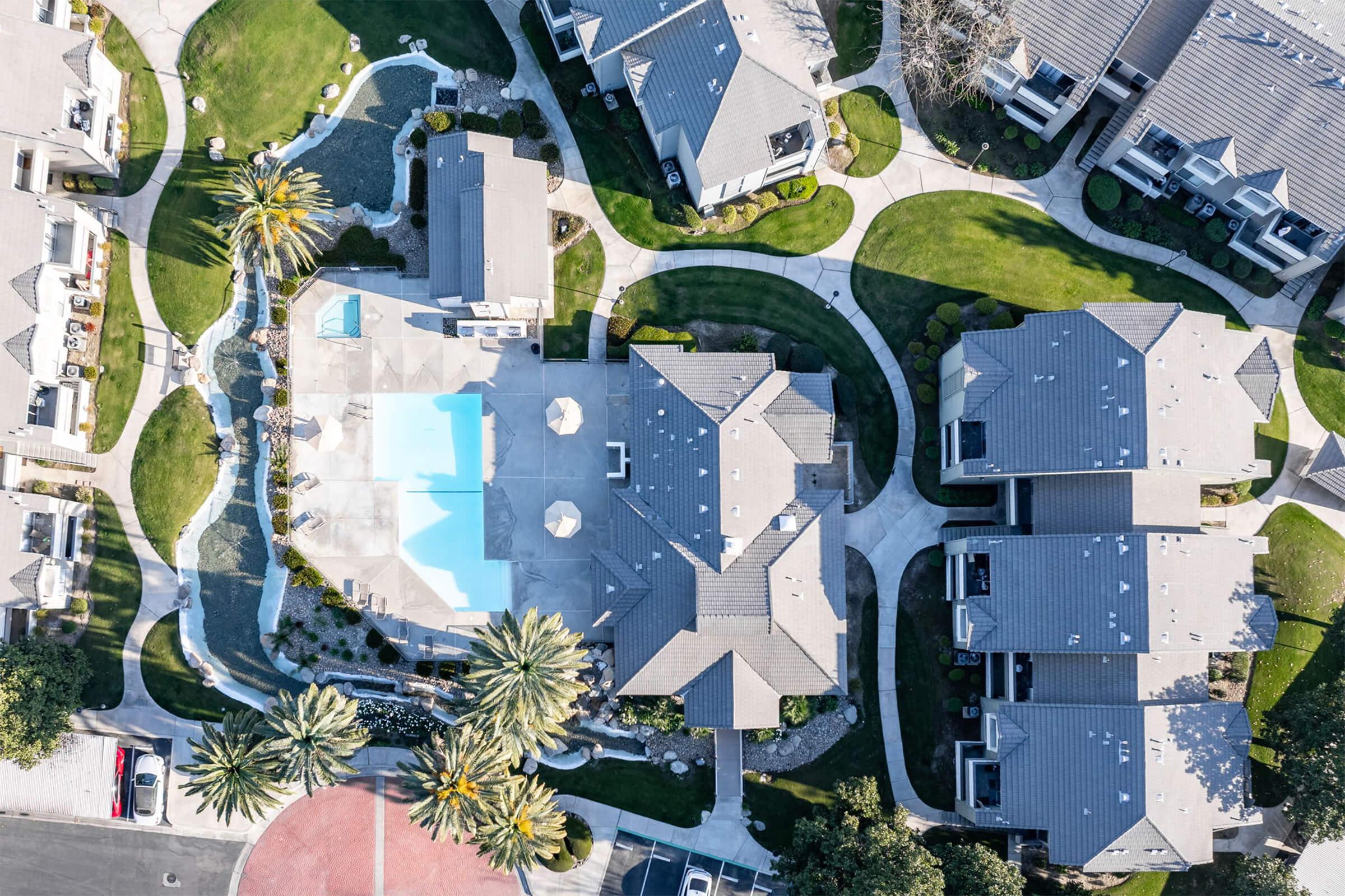 Aerial view of a residential community featuring several houses and a central pool area. The landscape includes manicured lawns, palm trees, and curved pathways connecting the buildings. The pool is blue with lounge chairs arranged around it, providing a relaxing atmosphere in a well-maintained environment.