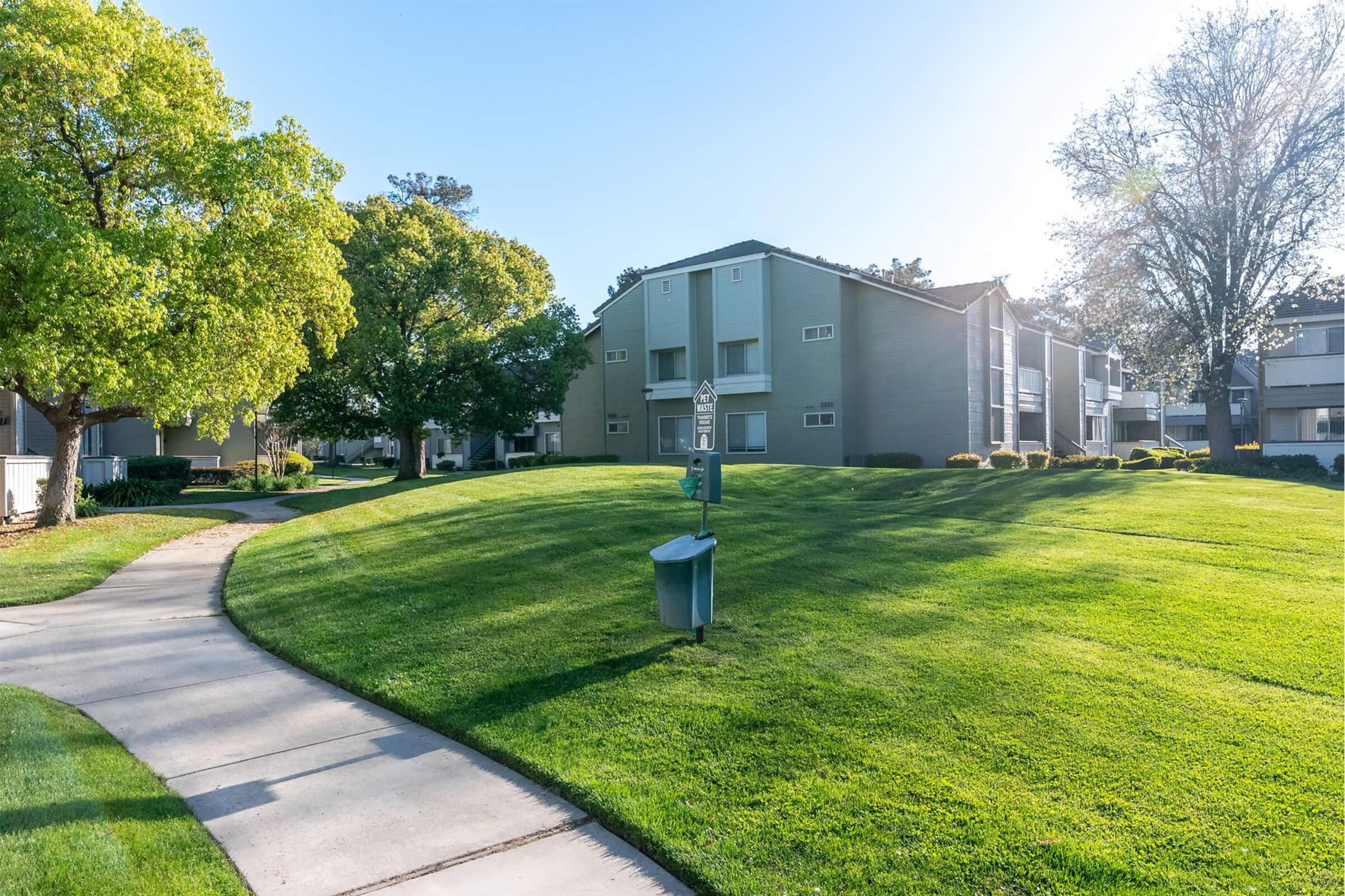 A sunny view of a well-maintained apartment complex surrounded by green lawns and trees. A curved walkway leads through the landscape, with a mailbox visible along the path. The scene conveys a peaceful and inviting residential atmosphere.
