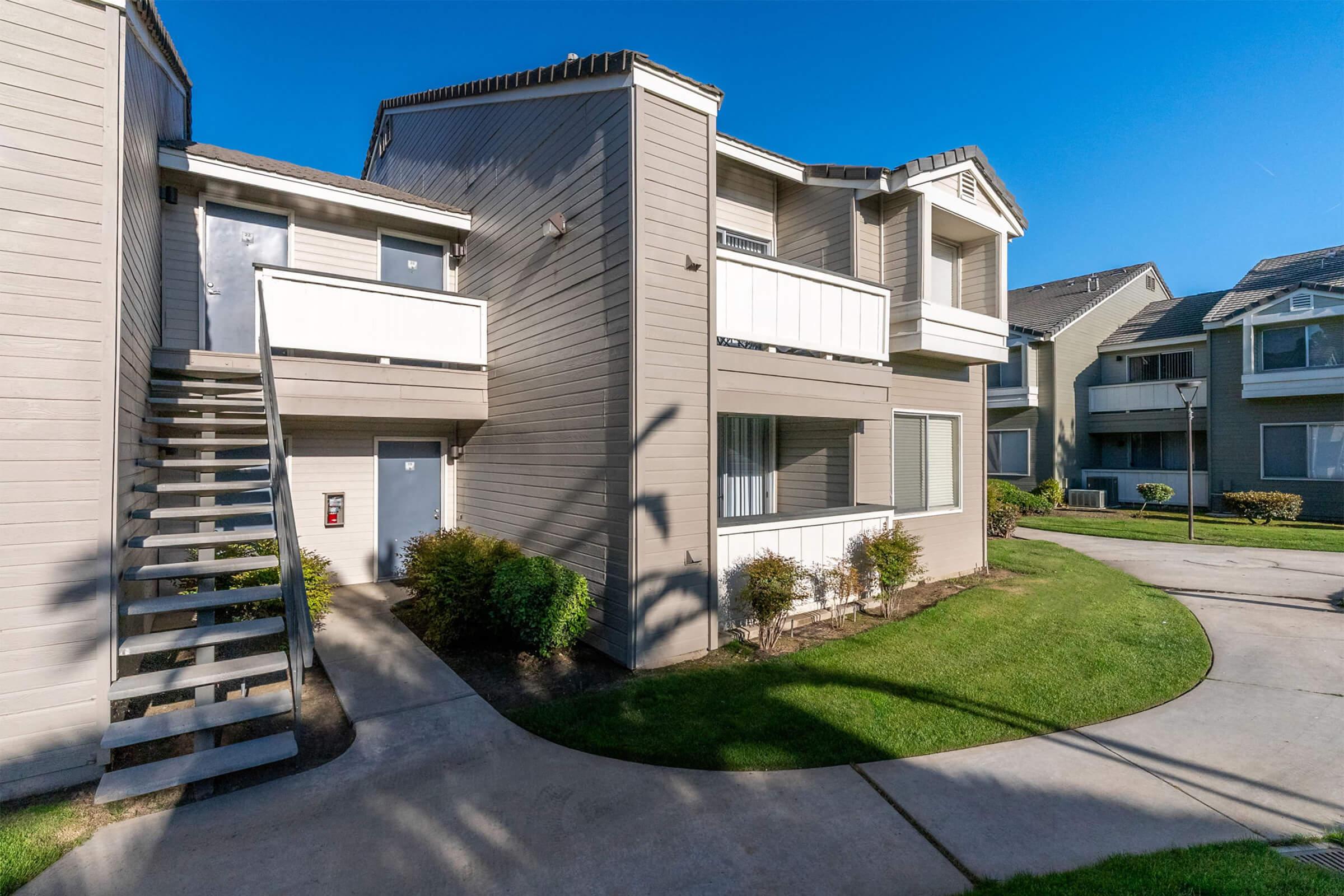 A two-story apartment building with beige siding, featuring a staircase leading to an upper unit. The landscape includes green grass and small shrubs, with additional units visible in the background under a clear blue sky.
