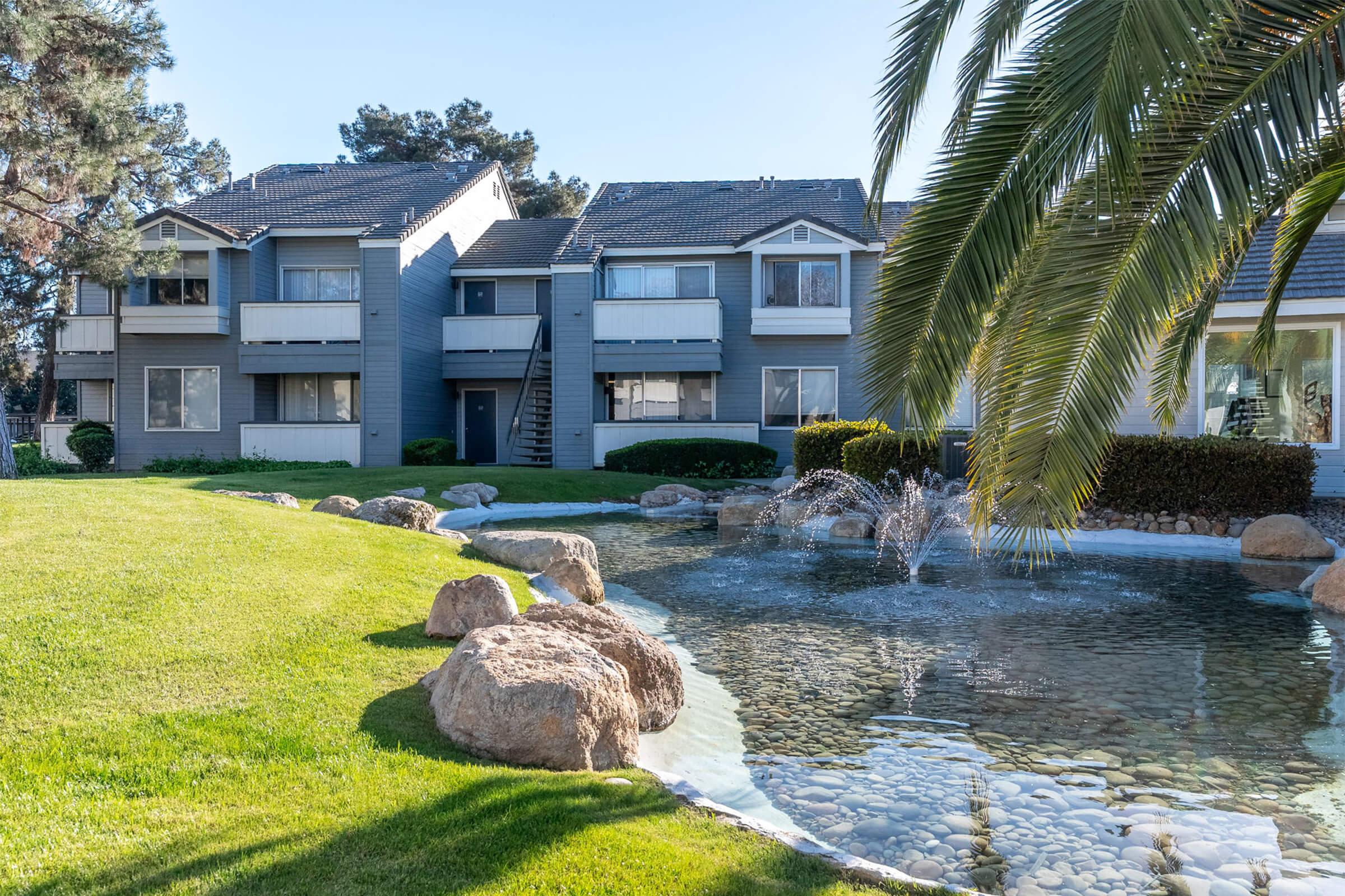 A peaceful residential complex featuring several two-story buildings surrounded by lush green grass, decorative boulders, and a small pond with a fountain in the foreground. Palm trees add to the serene atmosphere, creating an inviting outdoor space.