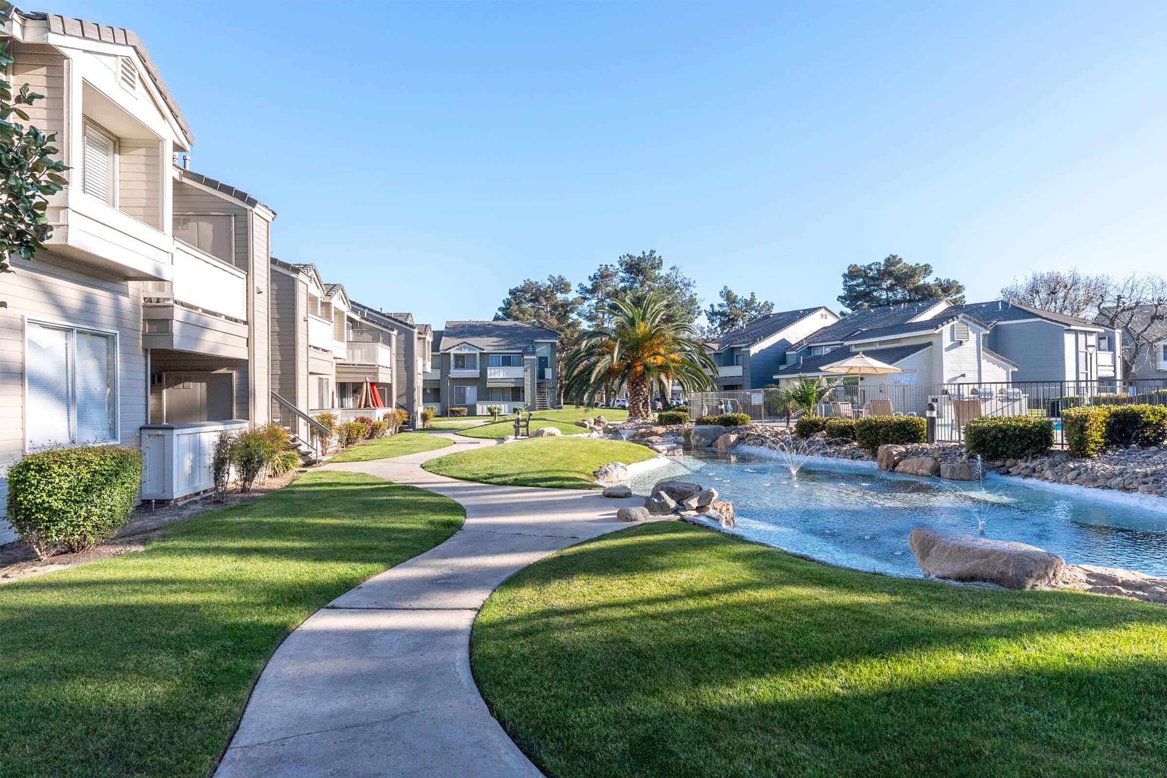 A landscaped pathway meanders through an apartment complex featuring lush green lawns, ornamental bushes, and a serene water feature. The scene includes modern buildings, palm trees, and a clear blue sky, creating a tranquil residential environment.