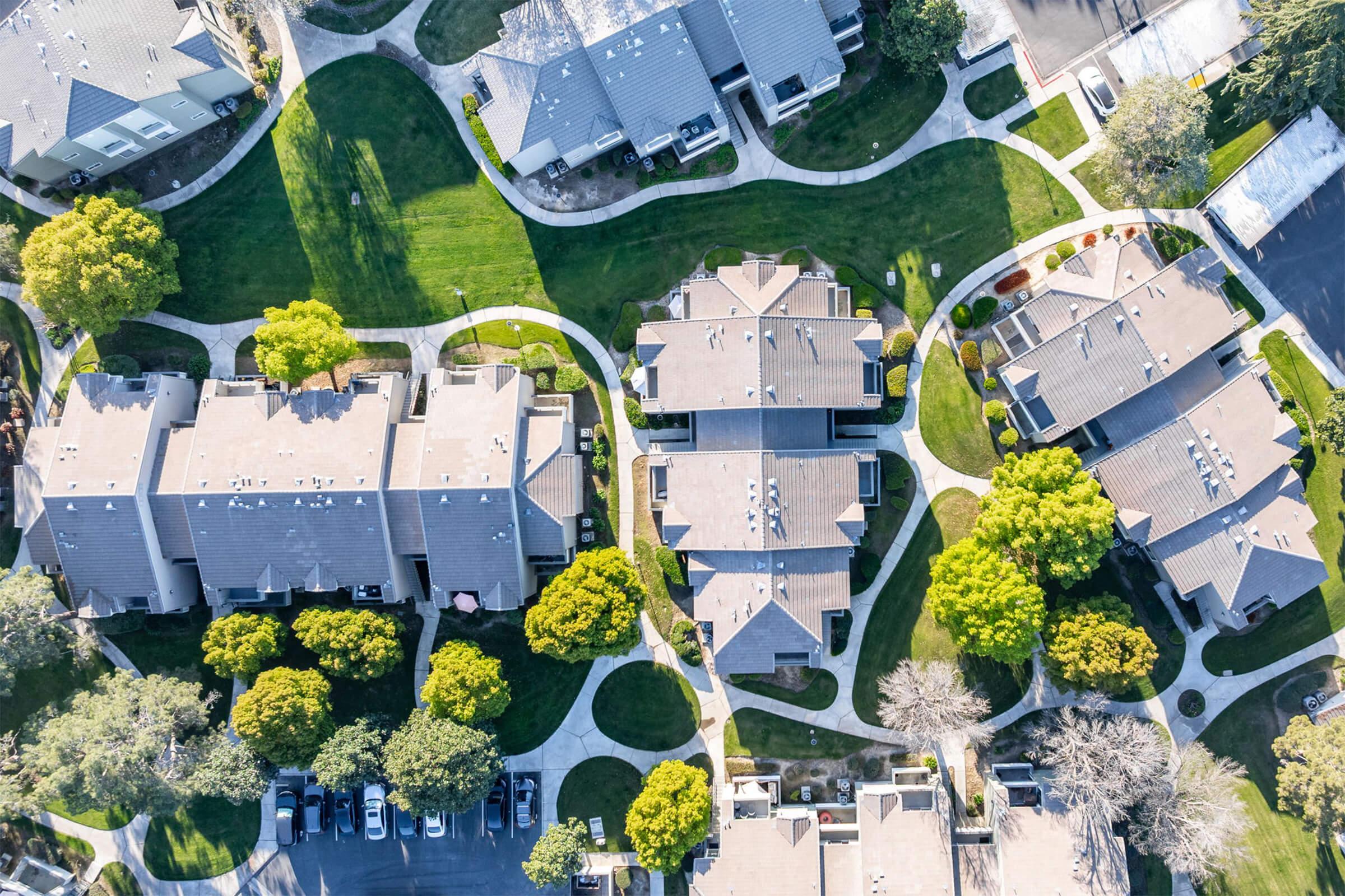 Aerial view of a residential neighborhood featuring several houses arranged in a circular layout, green lawns, winding pathways, and trees. The scene shows a well-maintained area with cars parked along the streets and walking paths connecting the homes.