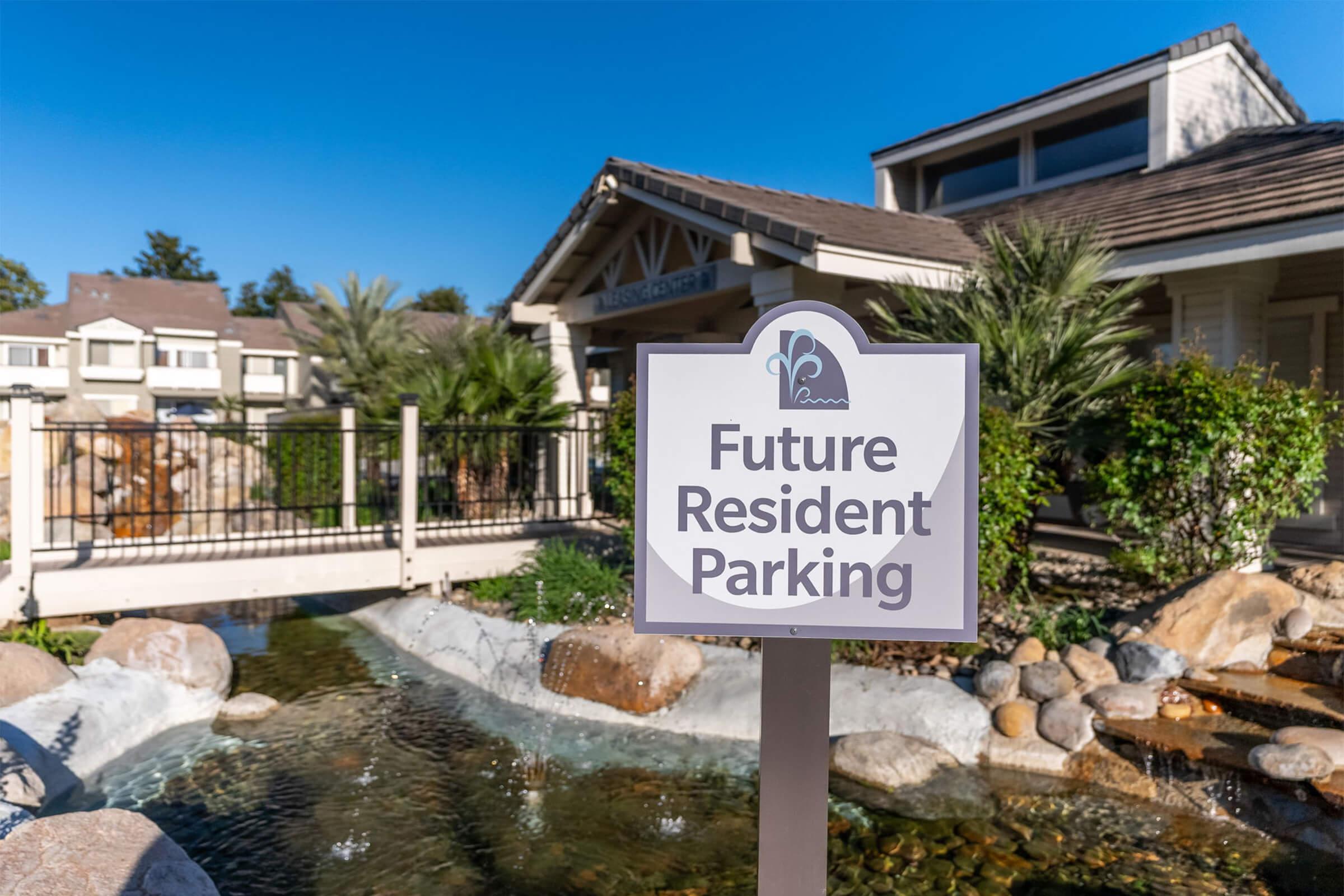 Sign indicating "Future Resident Parking" in front of a residential building, with a small pond and landscaped area featuring rocks and palm trees in the background under a clear blue sky.