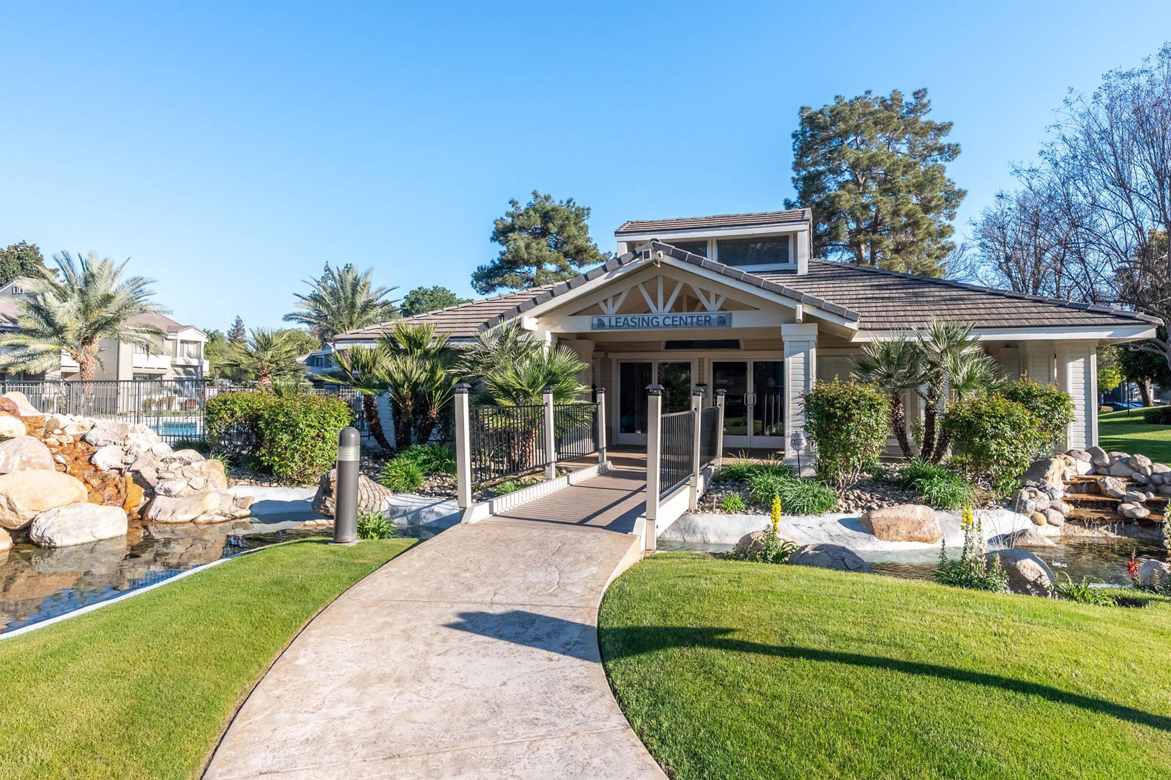A welcoming entrance to a building labeled "Inspiring Center," featuring a stone pathway leading to the door, surrounded by lush greenery, palm trees, and rocks. A serene water feature is visible on the left, with clear blue skies above, creating a bright and inviting atmosphere.
