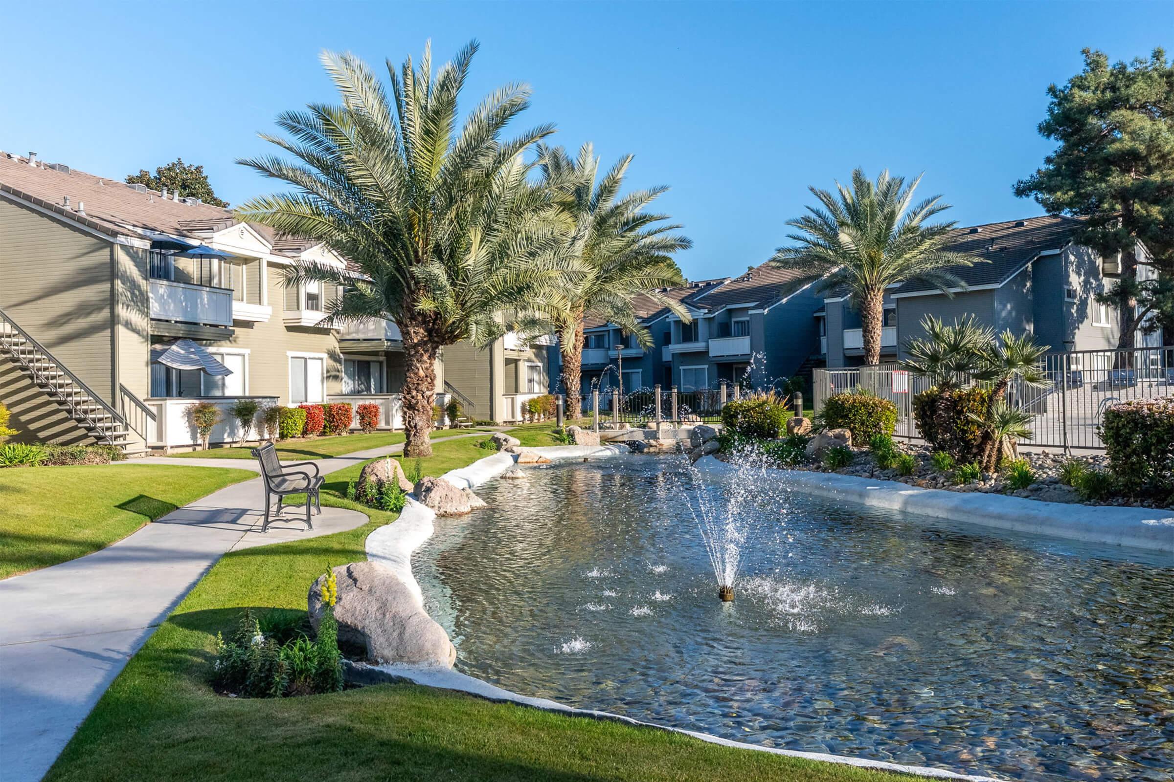 A serene landscaped area featuring palm trees, a small pond with a fountain, and neatly maintained grass. Surrounding the pond are well-kept pathways and benches, with two-story apartment buildings visible in the background under a clear blue sky.