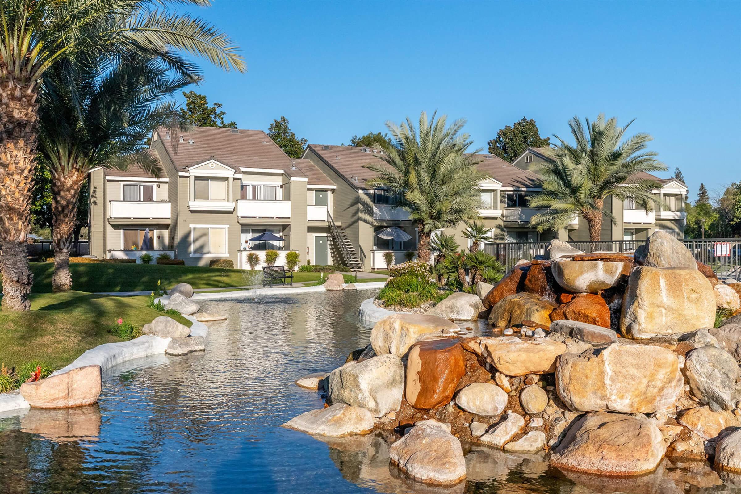 A tranquil scene featuring a landscaped area with a pond and a rock formation in the foreground. In the background, there are multi-story residential buildings surrounded by palm trees and greenery, set against a clear blue sky. The setting conveys a serene and inviting atmosphere.