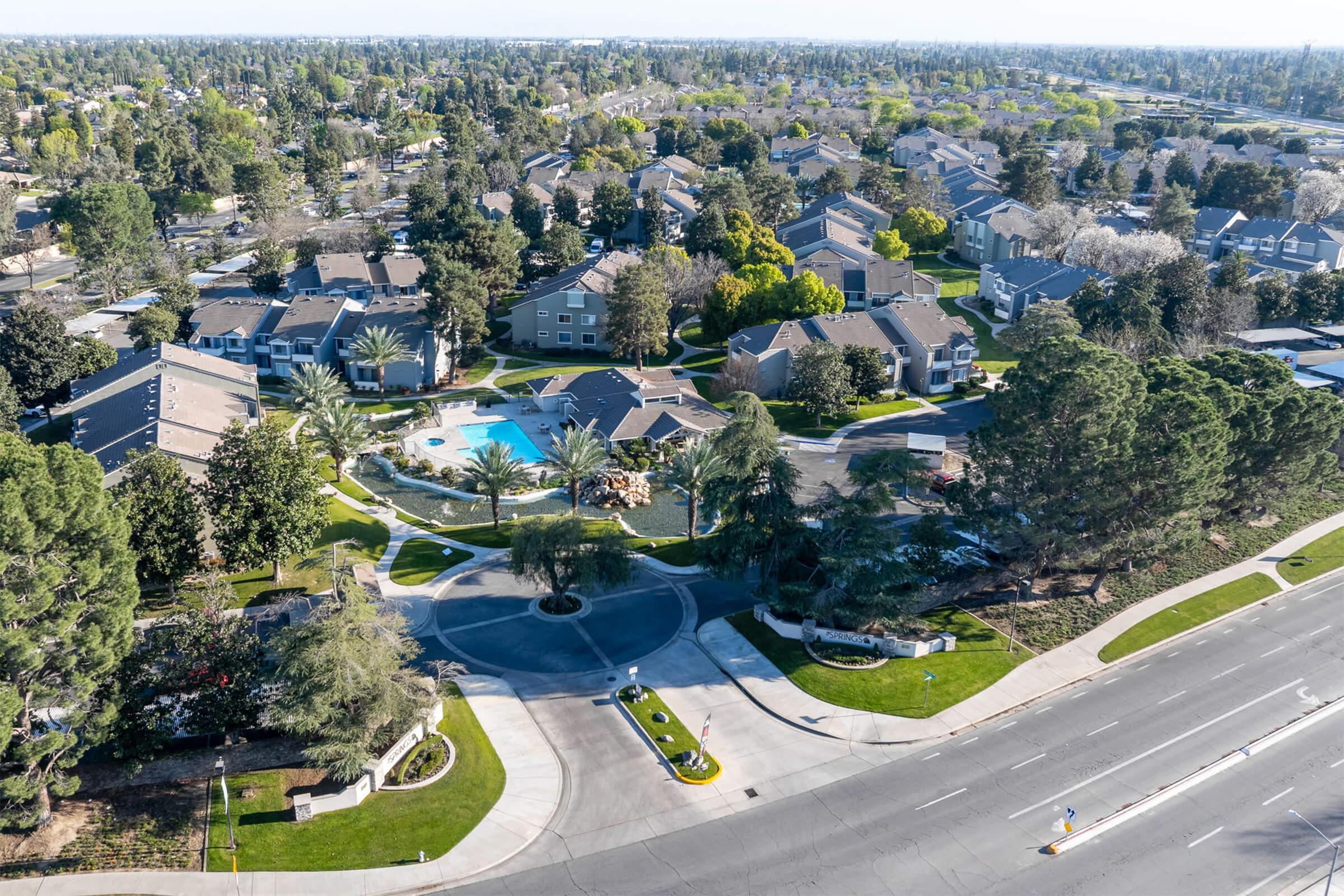 Aerial view of a residential neighborhood featuring multiple apartment buildings, a central pool area, and landscaped greenery. The layout includes roads, trees, and lush gardens, showcasing a suburban environment with a mix of community amenities and housing.
