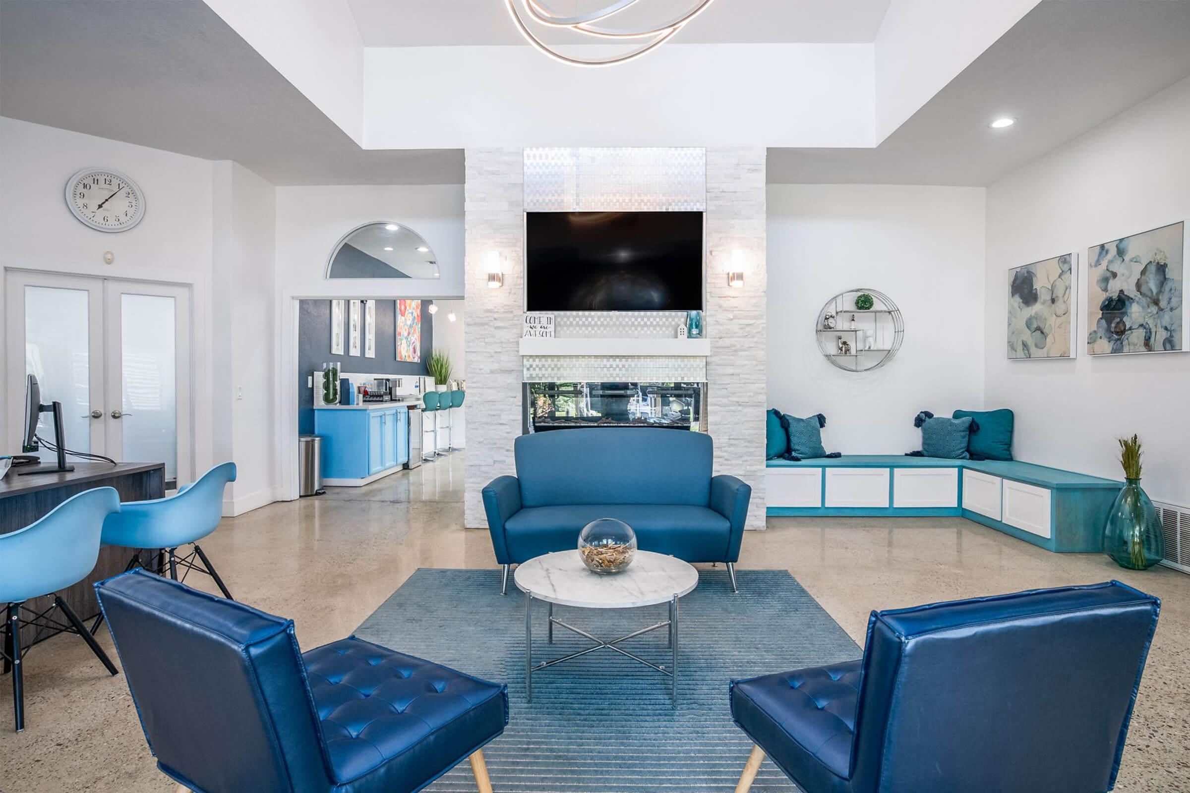 Modern living room with blue furniture, featuring a stylish couch and armchairs, a round coffee table, and a decorative wall with a fireplace. The space has neutral walls, a high ceiling, and contemporary decor, including artwork and a wall clock. Natural light brightens the area.