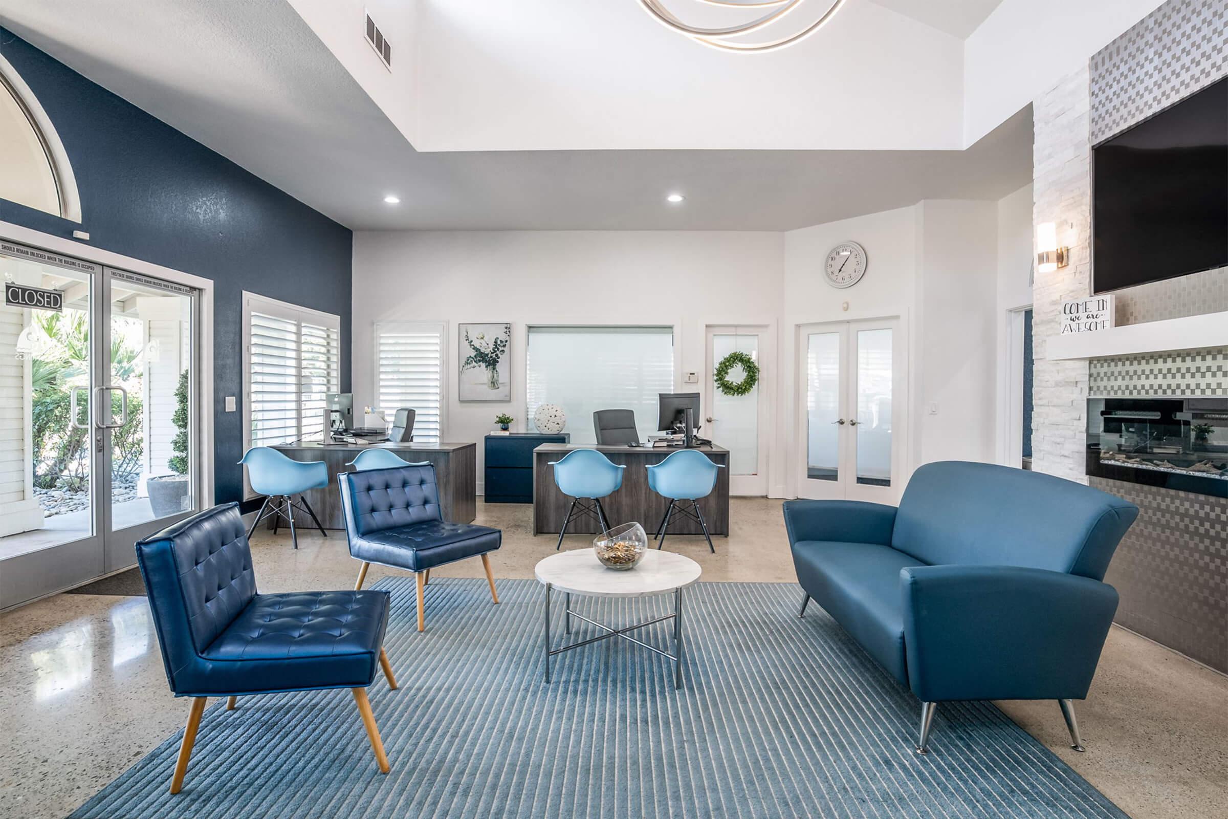 A modern reception area featuring stylish blue seating, a round coffee table, and a textured rug. Behind the seating, there are glass doors leading outside, and a front desk area with two workstations. The space has large windows and is well-lit with contemporary light fixtures.