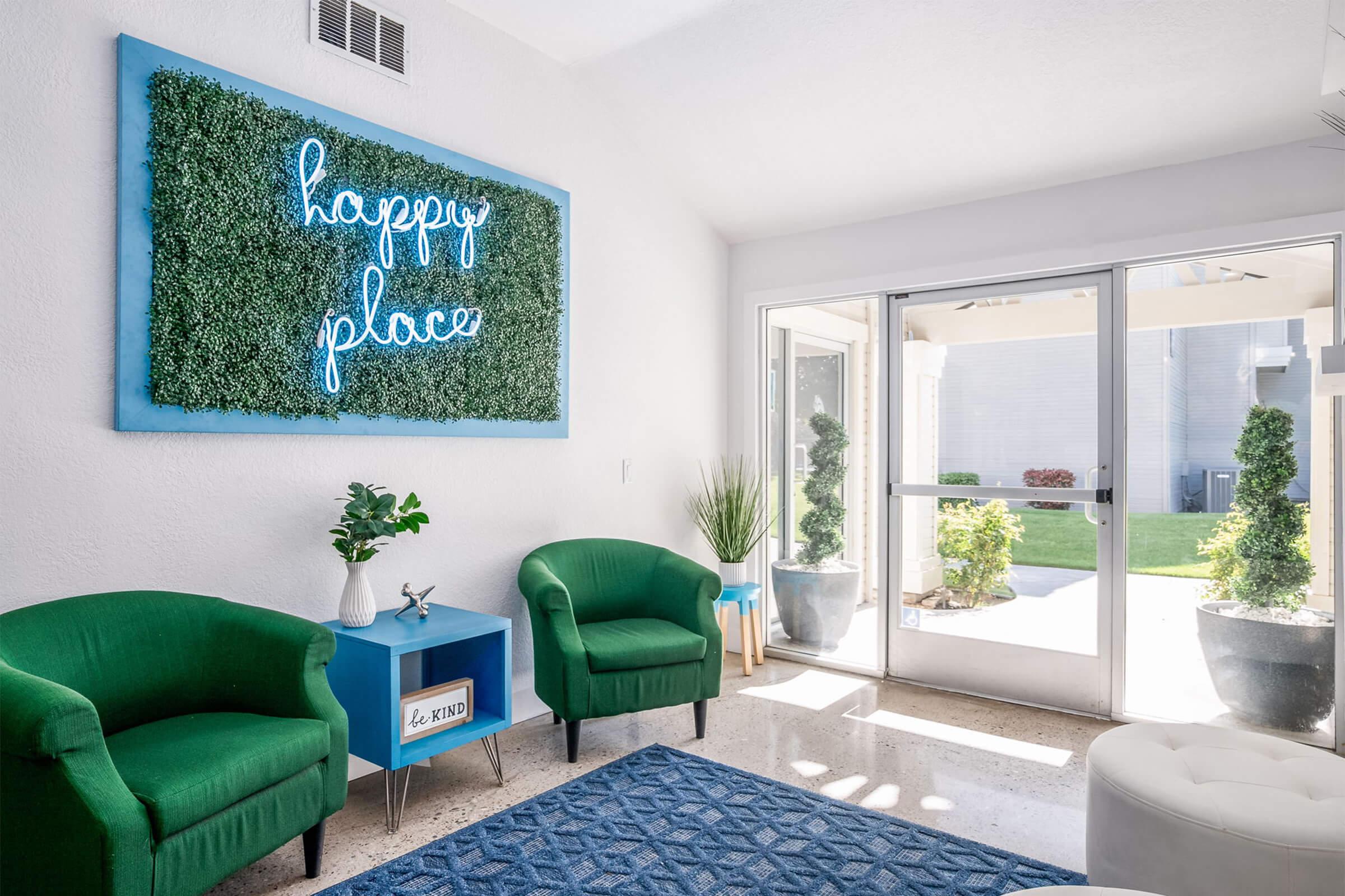A bright and inviting room featuring two green armchairs, a blue side table with a decorative item, and a large green wall art piece with the words "happy place" in neon. The room has large windows allowing natural light, with plants and an outdoor view visible. The decor is modern and cheerful.