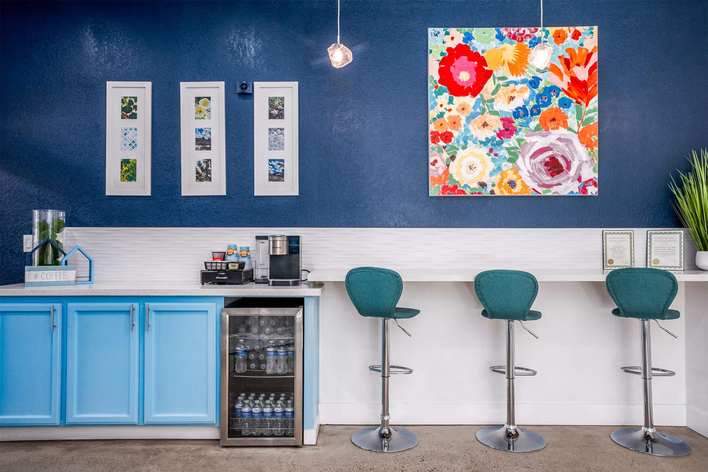 A bright and modern coffee station featuring a blue cabinetry, a coffee maker, and a mini refrigerator. Above the counter, colorful flower-themed artwork is displayed on the wall, alongside three framed pictures. The space includes stylish bar stools set against a white countertop, creating a welcoming atmosphere.