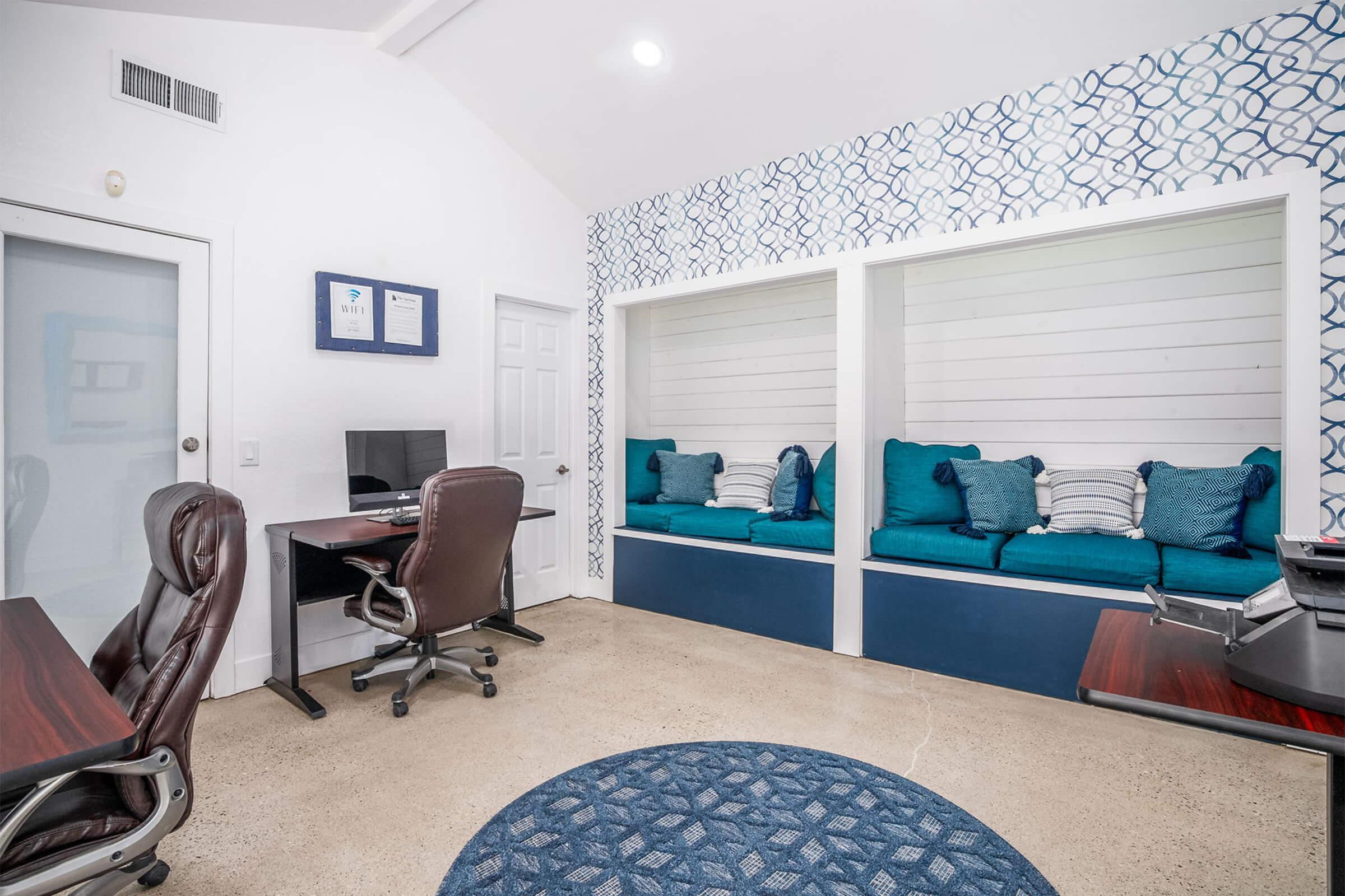 A modern office space featuring two work areas, each with a desk and an ergonomic chair. Soft blue cushions are positioned in cozy nooks along the wall. The decor includes a geometric patterned wallpaper and a round rug on a concrete floor, creating a bright and inviting atmosphere.