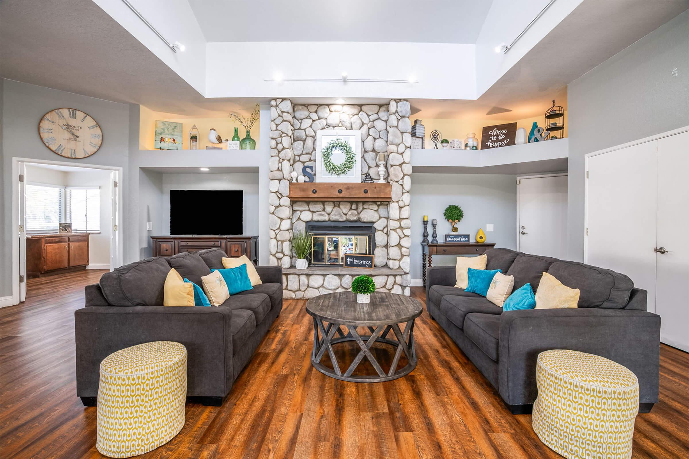 A modern living room featuring a stone fireplace as a focal point, flanked by two cozy gray sofas with colorful cushions. A round coffee table sits in the center, accompanied by decorative yellow poufs. Bright natural light fills the space, highlighting a peaceful and inviting atmosphere.