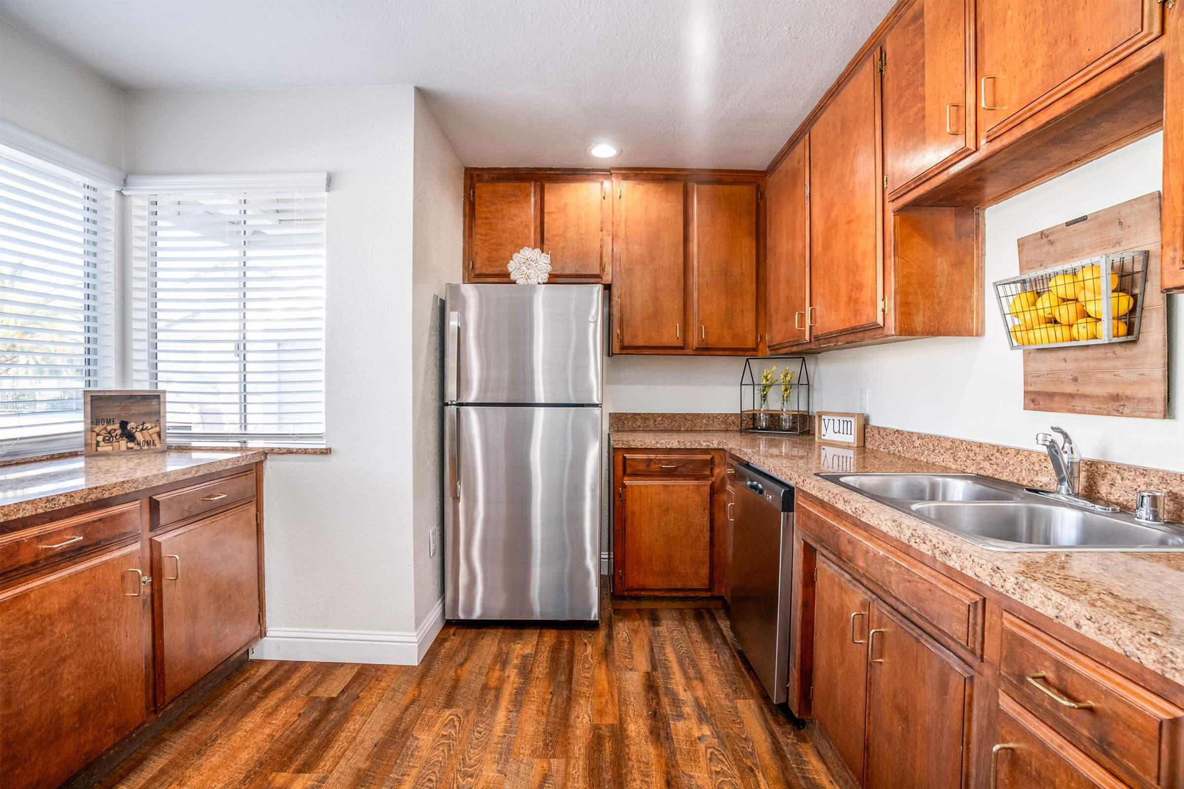 Bright and modern kitchen featuring wooden cabinets, stainless steel appliances including a refrigerator and dishwasher, and a countertop with a double sink. Natural light streams in through window blinds, enhancing the inviting atmosphere. Decor includes a basket of lemons and a small decorative item on the countertop.