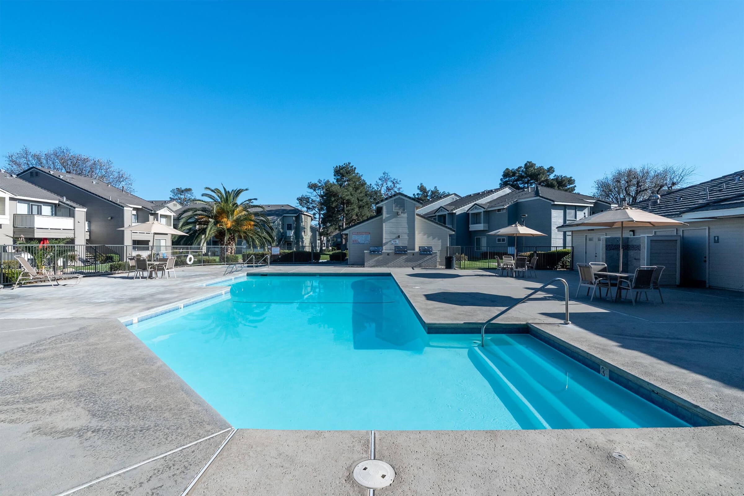 A clear blue swimming pool surrounded by a concrete deck, with lounge chairs and umbrellas nearby. Apartment buildings and palm trees in the background under a bright blue sky. A few shaded seating areas are visible, creating a relaxing outdoor space.