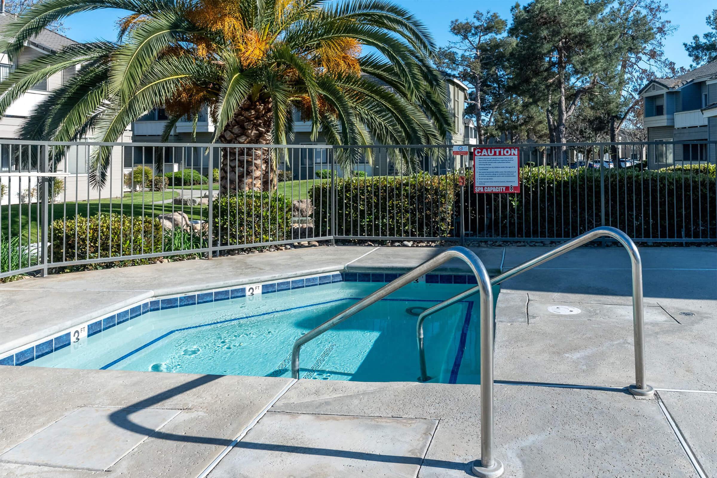 A clear view of a spa or hot tub with steps leading in, surrounded by a fenced area. In the background, a palm tree and landscaped shrubs are visible. A caution sign is posted nearby. The scene is set in a residential area with apartment buildings in the distance under a clear blue sky.